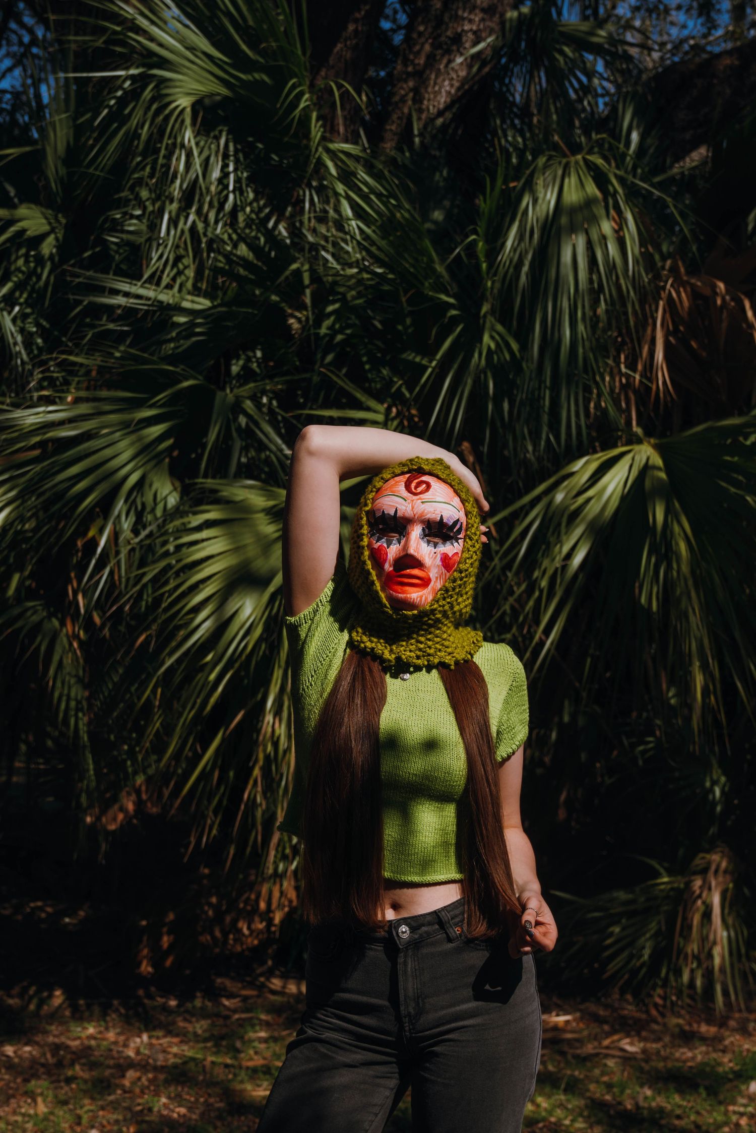 Woman in green knit crop top wearing a crocheted face mask with face paint among palm trees.