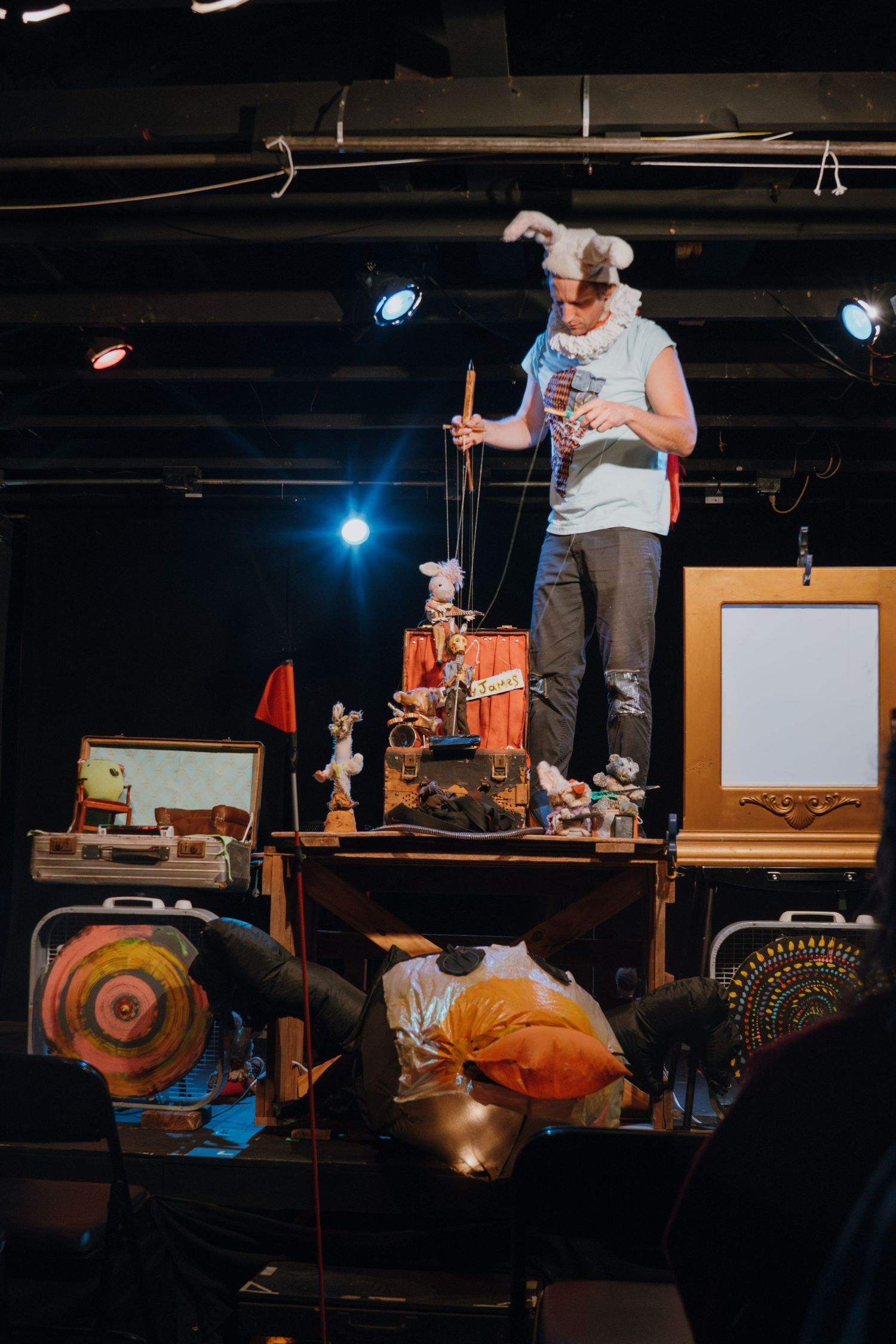Puppeteer performing on stage with marionette puppet among eclectic props under dramatic stage lighting.