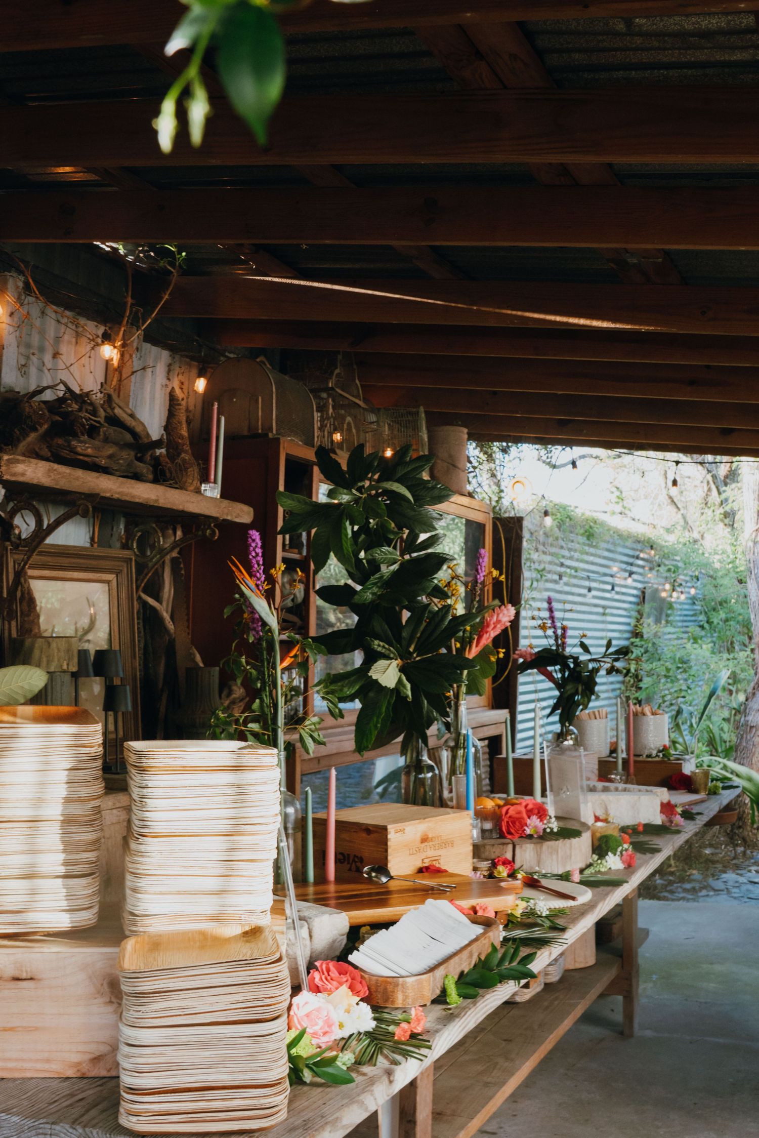 Rustic outdoor buffet table with stacked plates and tropical floral arrangements under string lights.