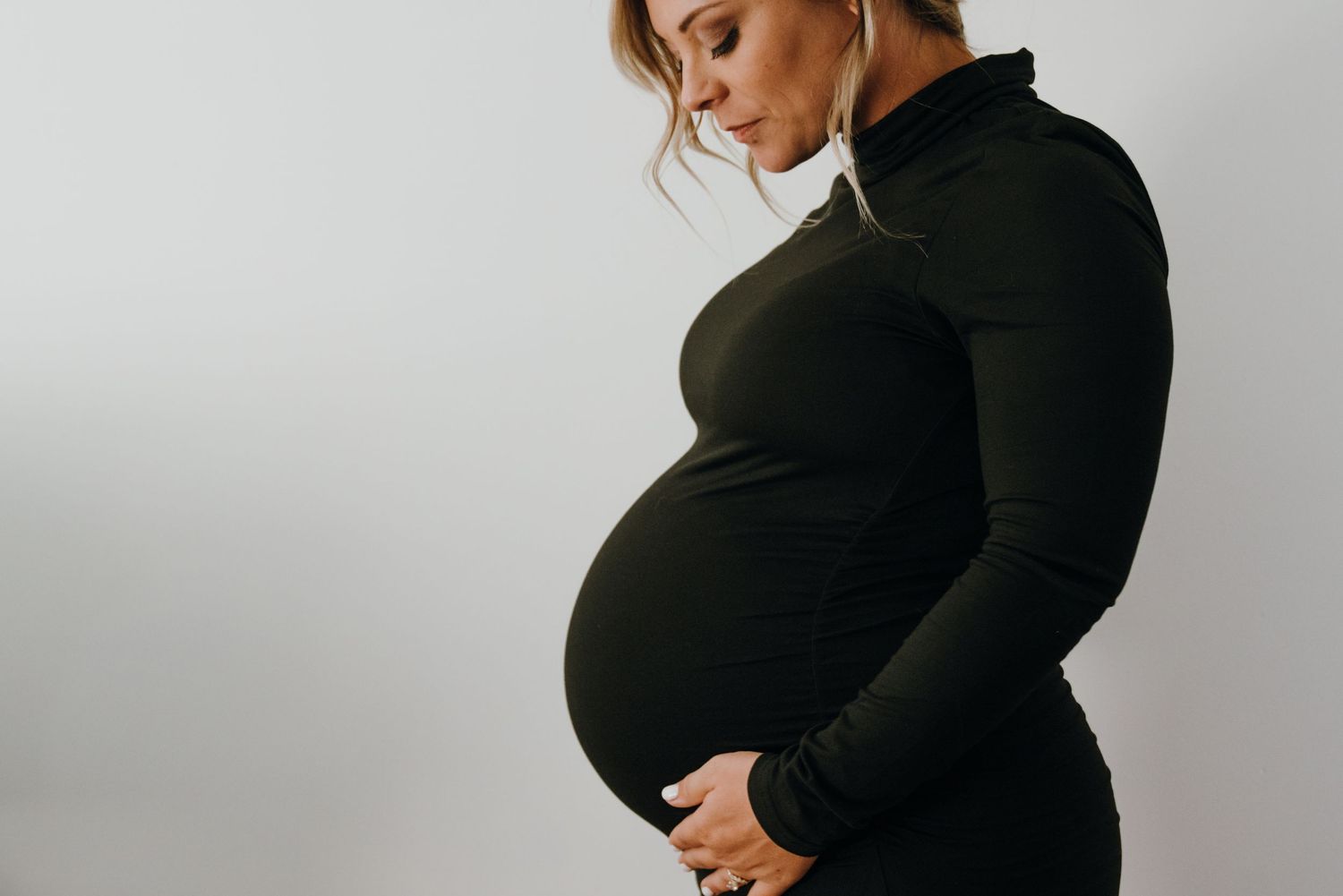 Pregnant woman in black long-sleeve dress gently holding her baby bump, side view.