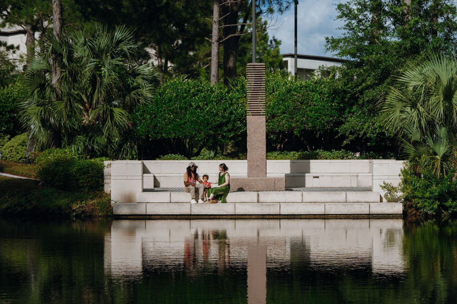 A family sits together on a white stone wall beside a calm reflective pond surrounded by lush greenery.