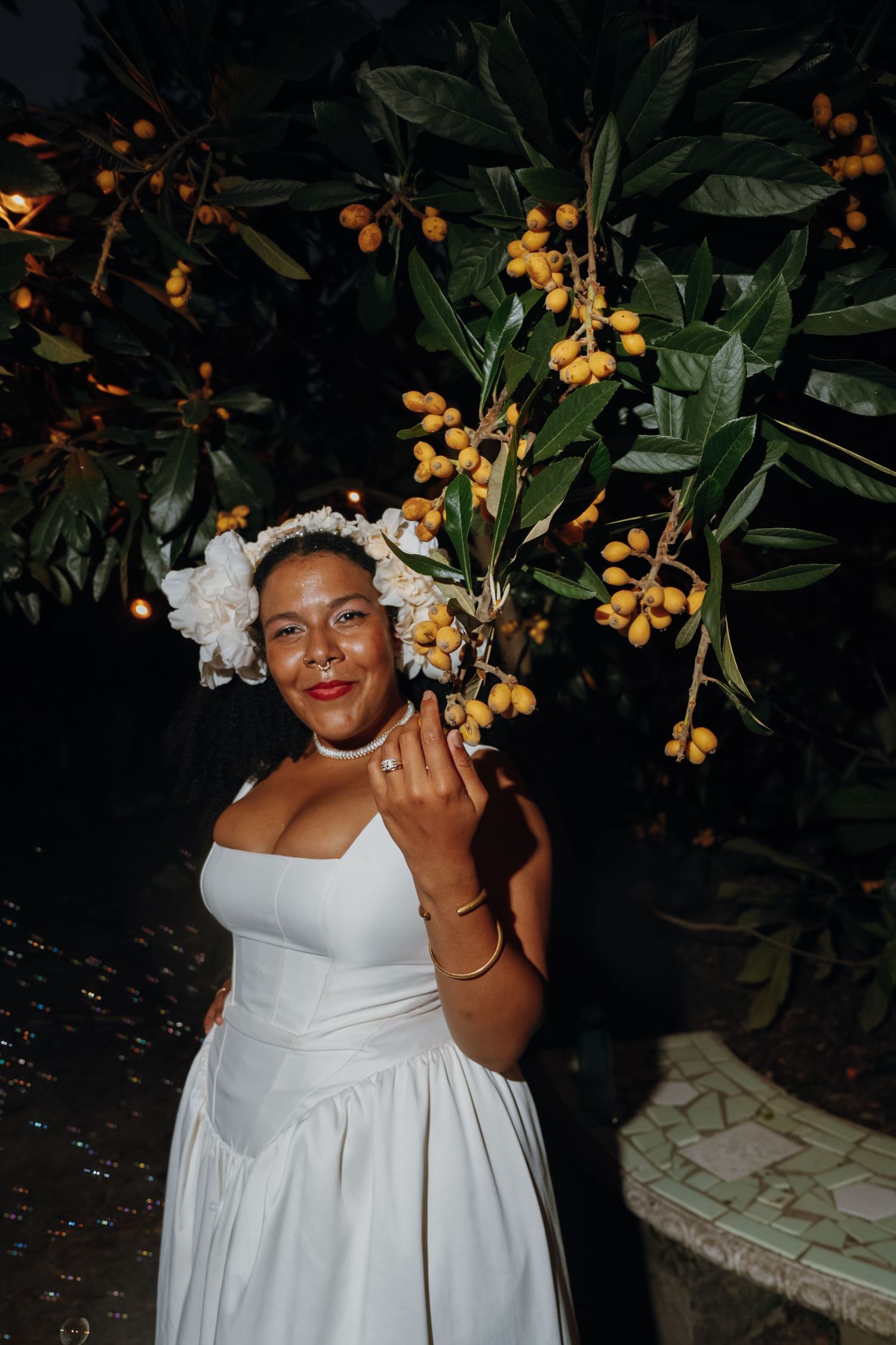 Bride in white strapless dress wearing floral crown holds yellow fruit from tree at night.
