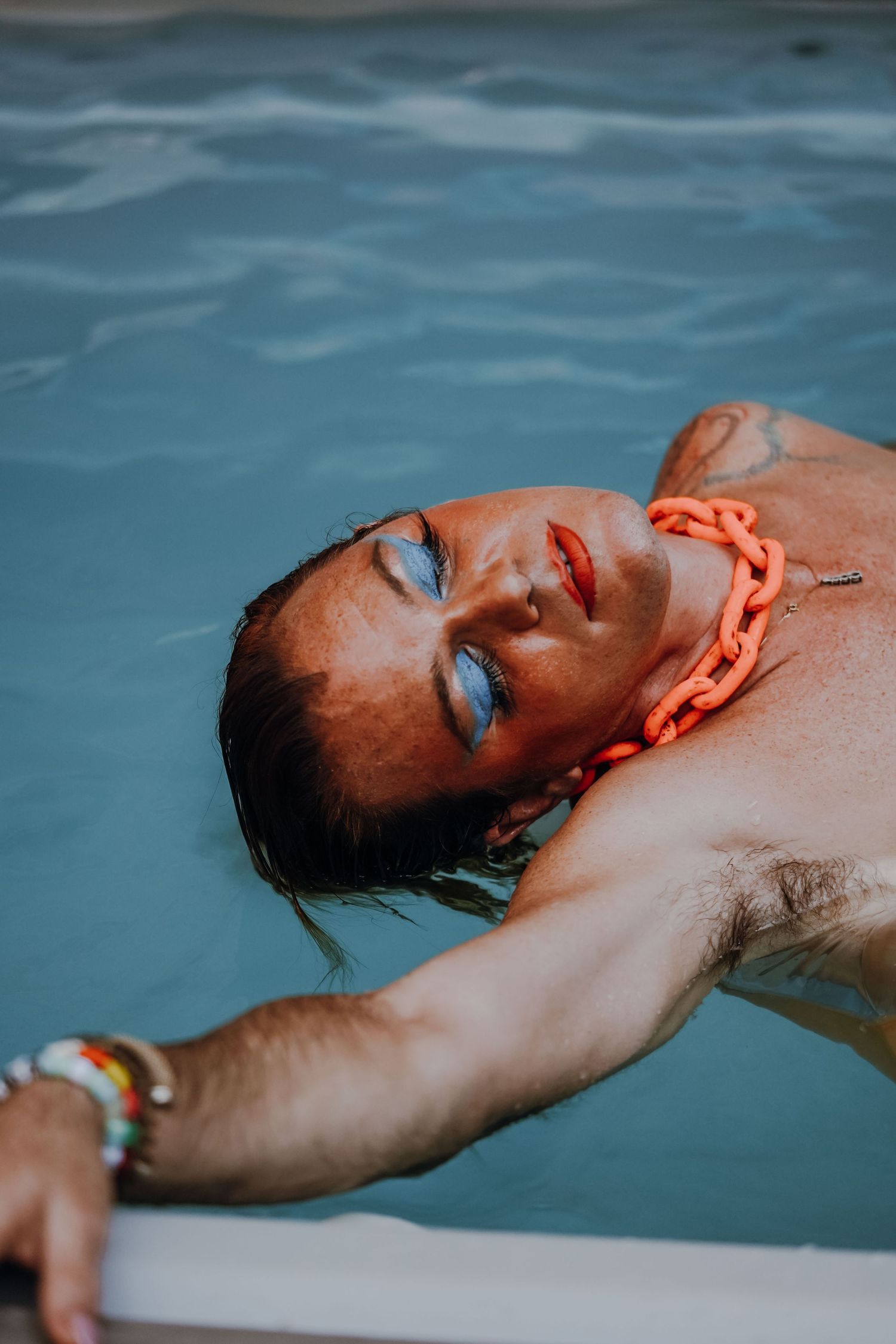 Man with blue eyeshadow and orange chain necklace relaxing in a swimming pool.