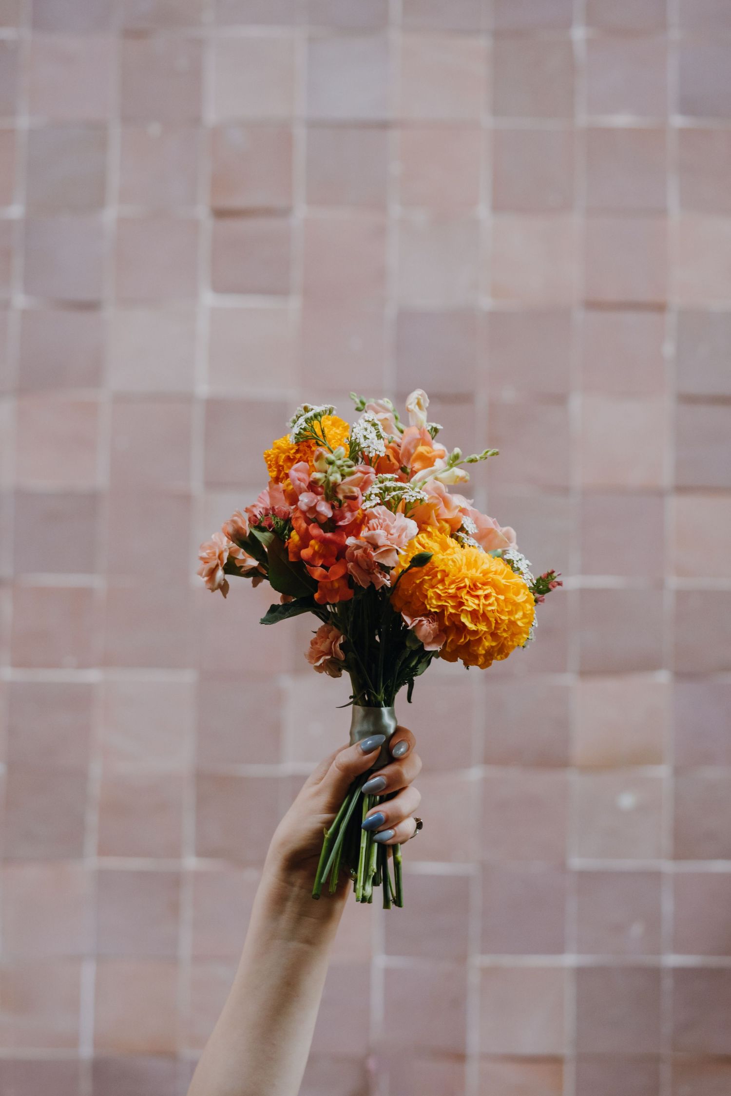 Hand holding a small bouquet of orange marigolds and pink flowers against a pink square tile wall.