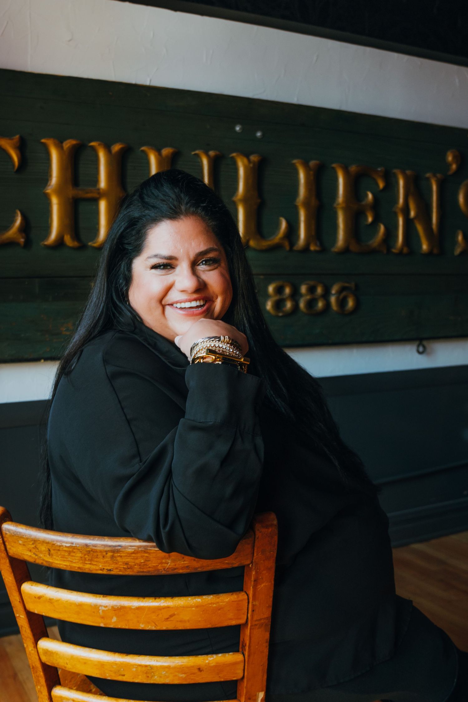 Smiling woman in black outfit seated at a wooden chair inside a vintage restaurant with antique signage.