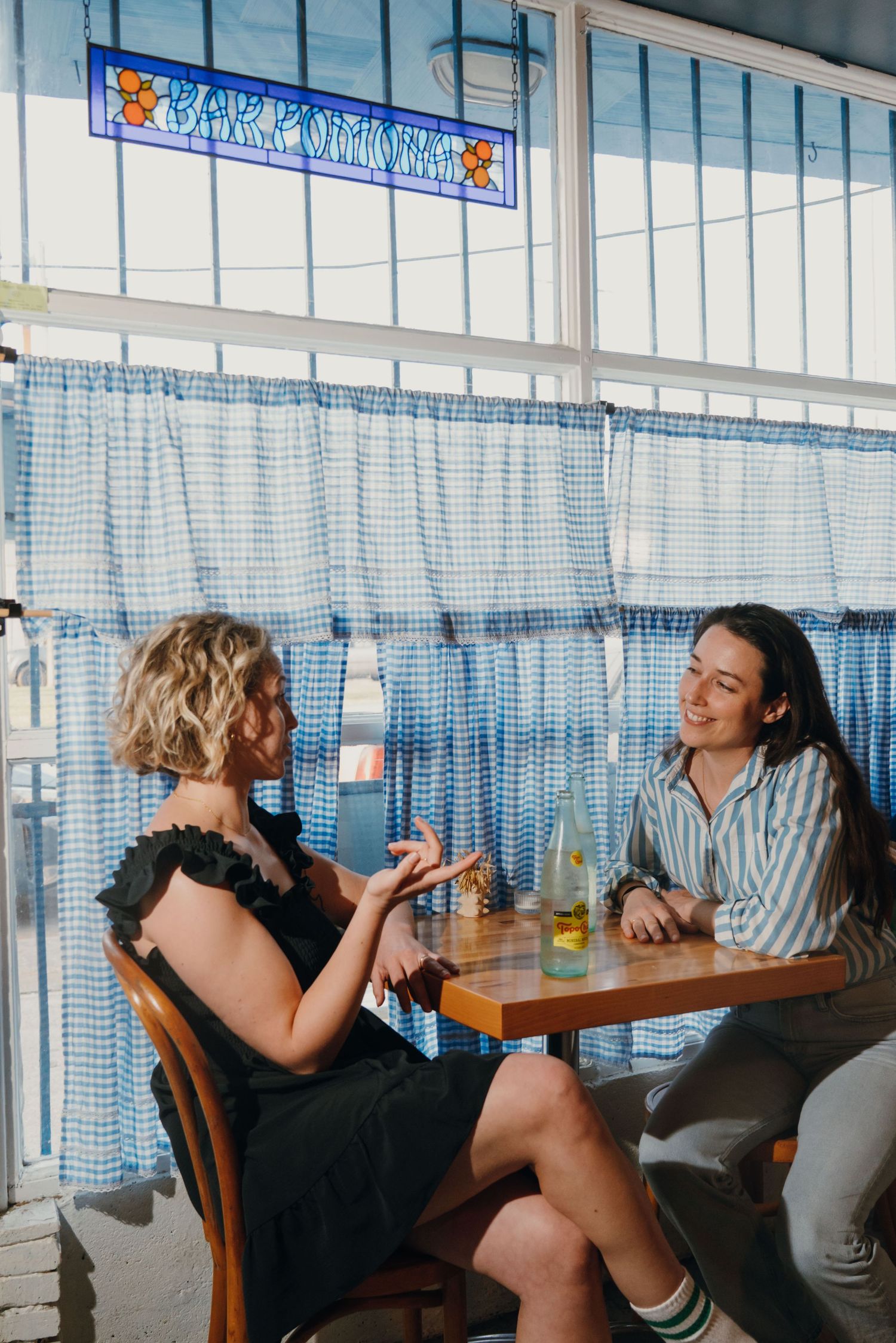 Two women enjoying conversation at a sunny restaurant booth with blue gingham curtains and drinks.