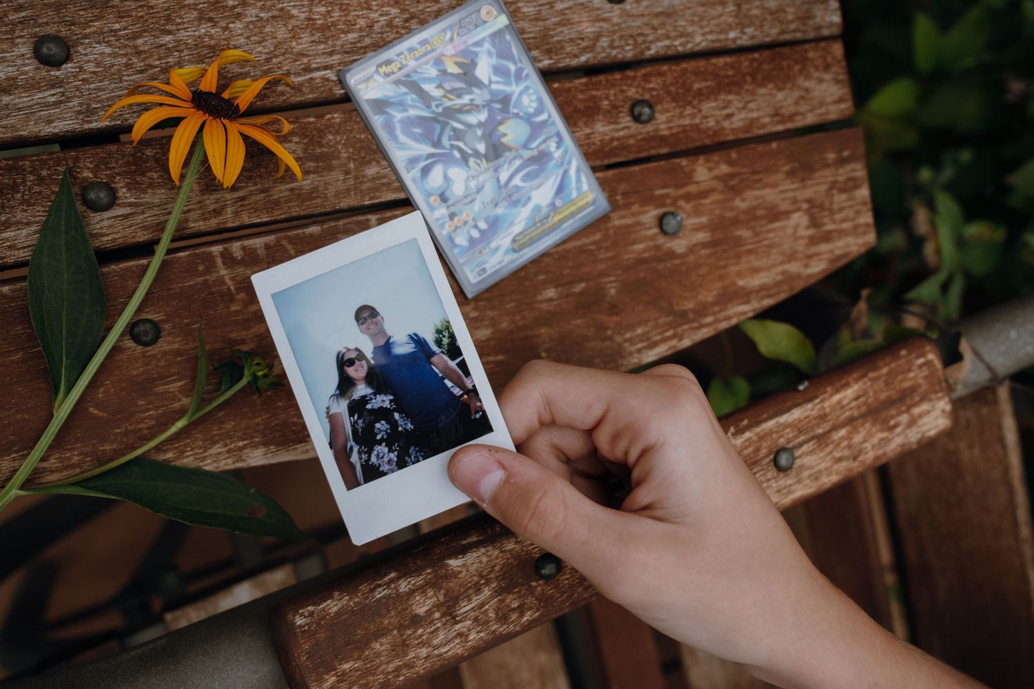 A hand holds a Polaroid photo of a family near a yellow flower and trinket on a rustic wooden bench.