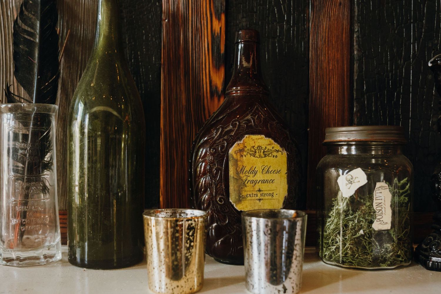 Collection of vintage bottles, mercury glass shot cups, and herb-filled mason jar on a wooden shelf.