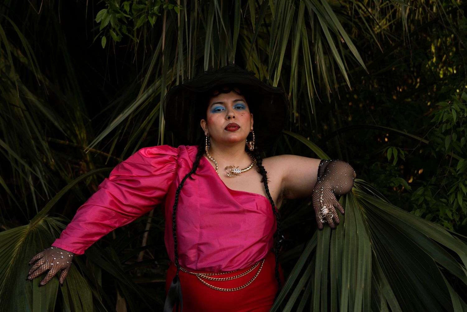 Woman in pink jacket and red skirt with black hat posing among lush tropical green leaves.