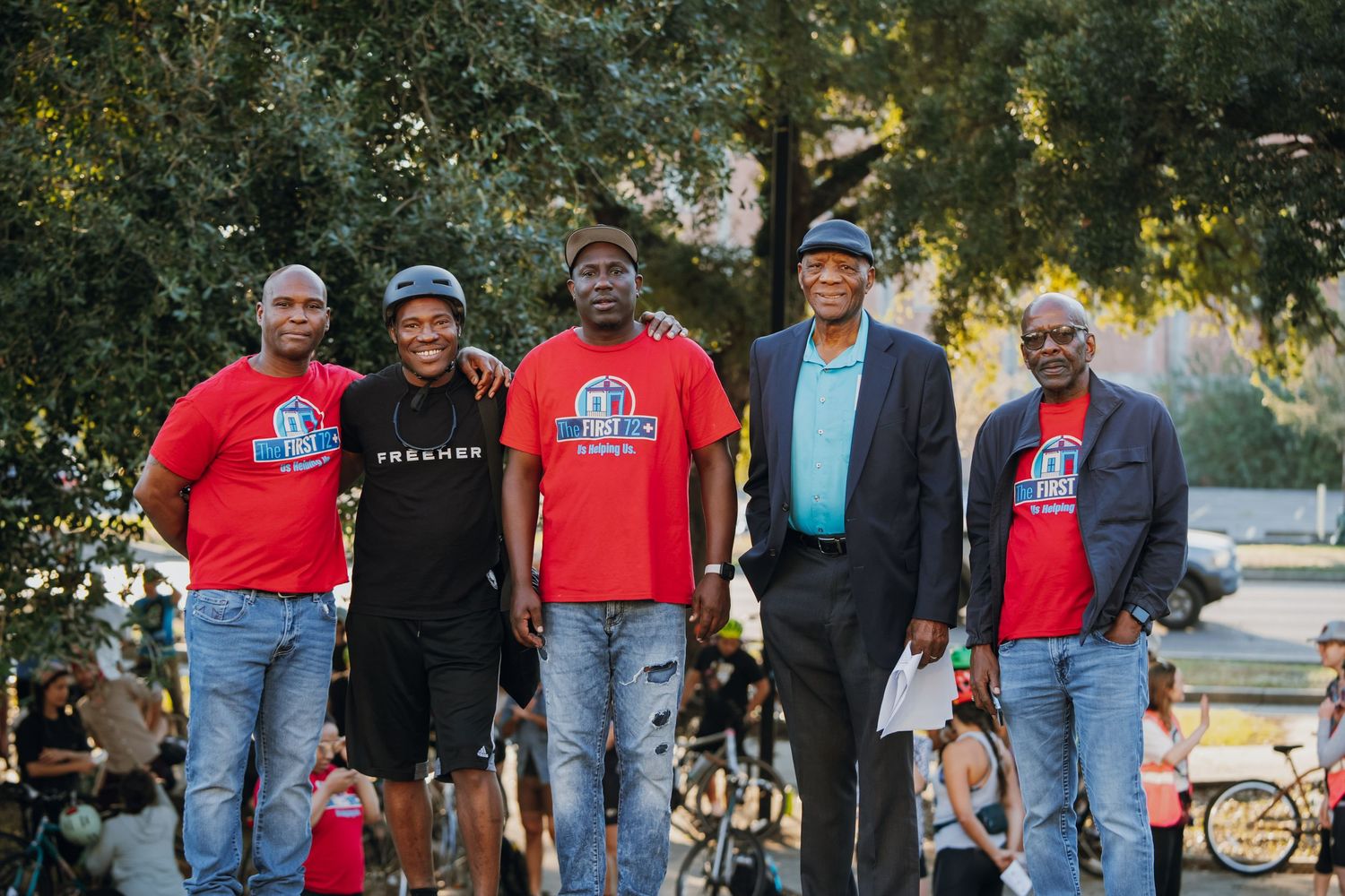 Five men in matching red shirts posing together outdoors under trees at community event.