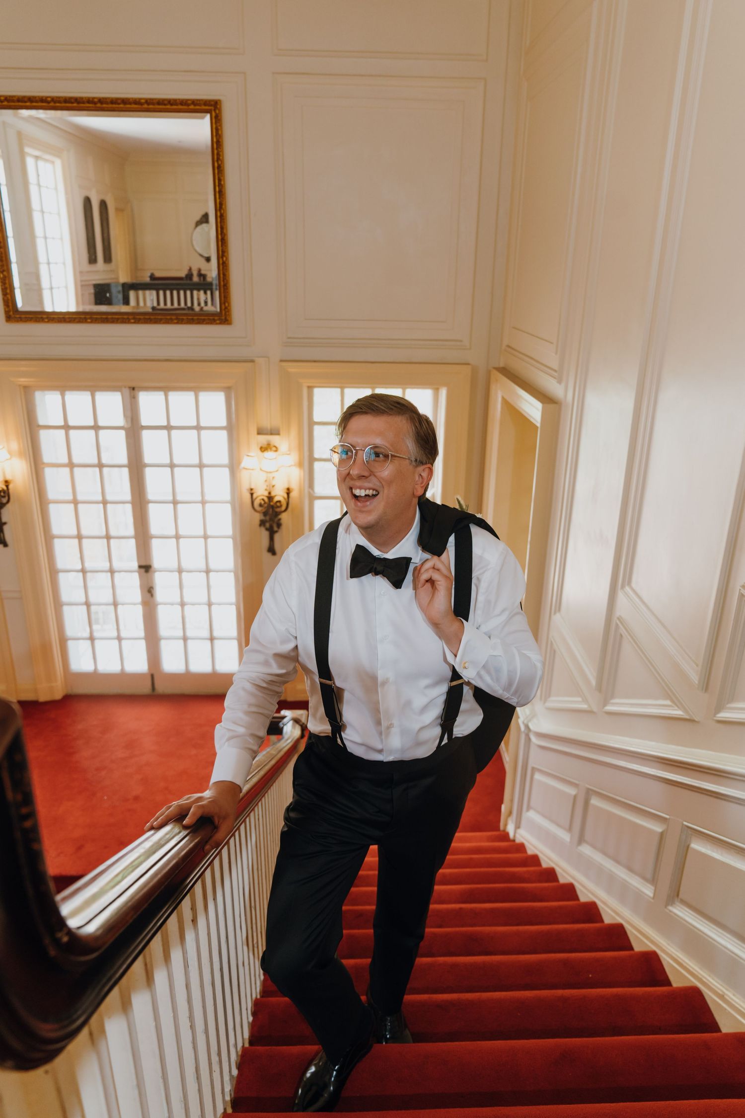 Happy groom in tuxedo with suspenders and bow tie laughing while ascending a red-carpeted staircase. New Orleans, Louisiana, Mississippi