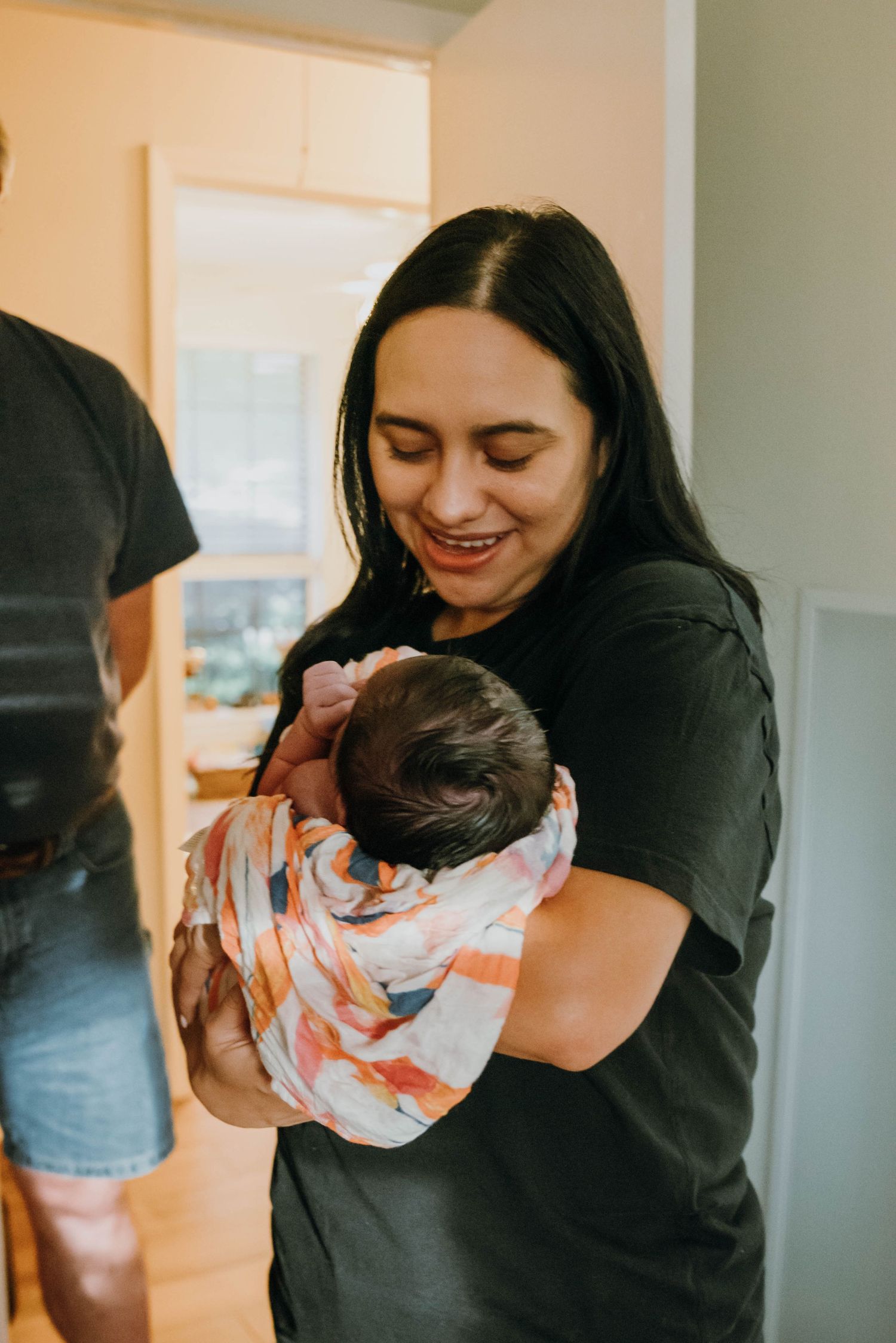 Smiling dark-haired woman in black shirt tenderly holding swaddled newborn baby indoors.