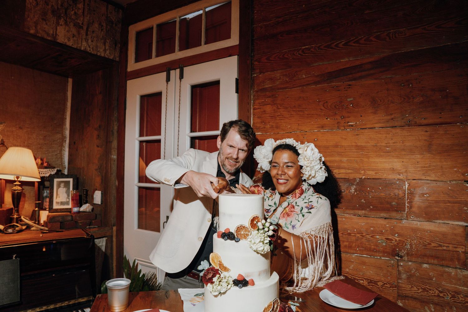 Smiling bride and groom cut their elegant white tiered wedding cake decorated with fresh fruit.