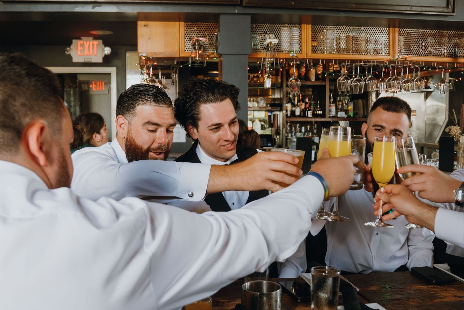 Groomsmen toasting with mimosas at a bar, smiling and celebrating before the wedding ceremony.