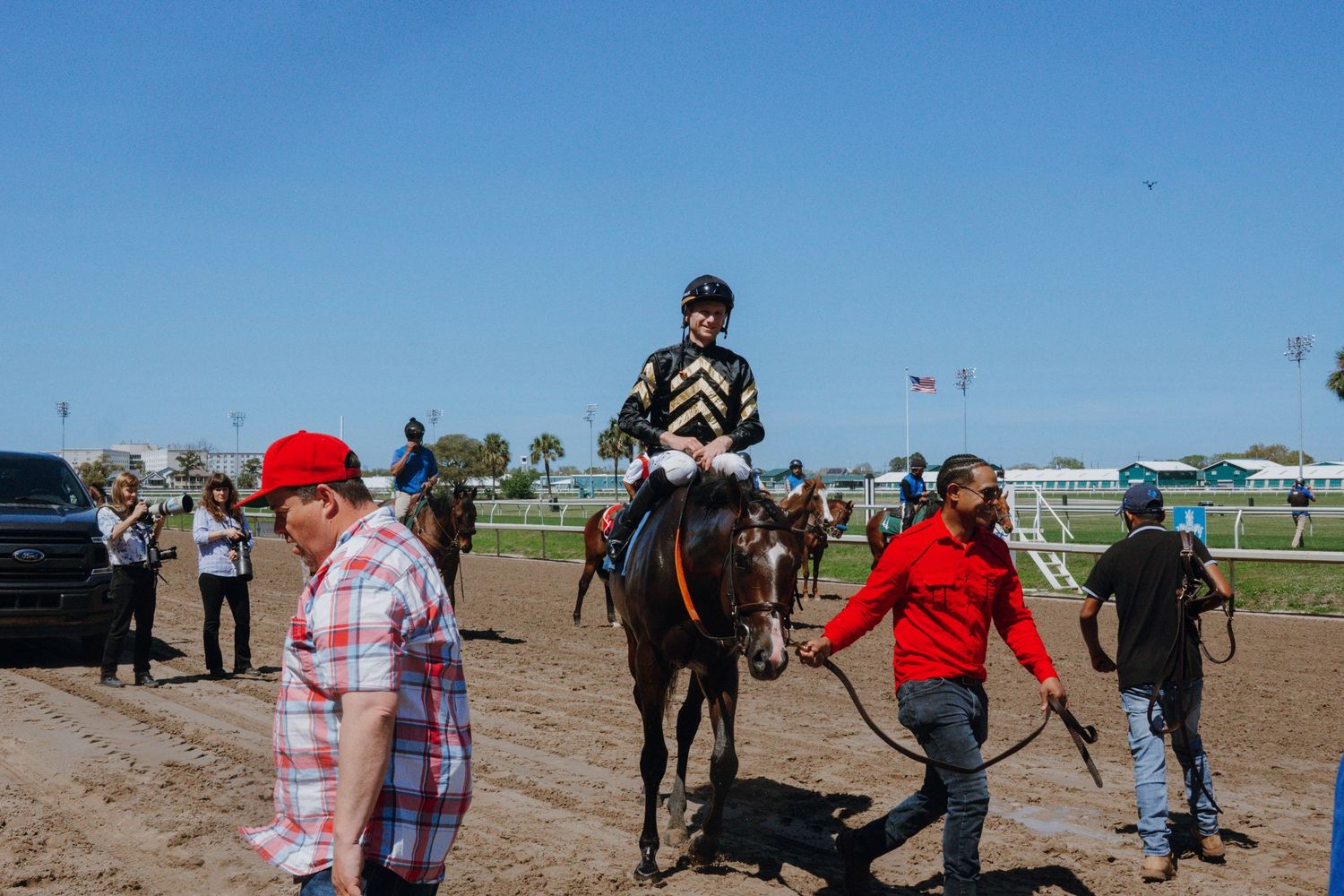 Jockey on brown horse at racetrack with handlers and spectators on sandy track under clear blue sky.