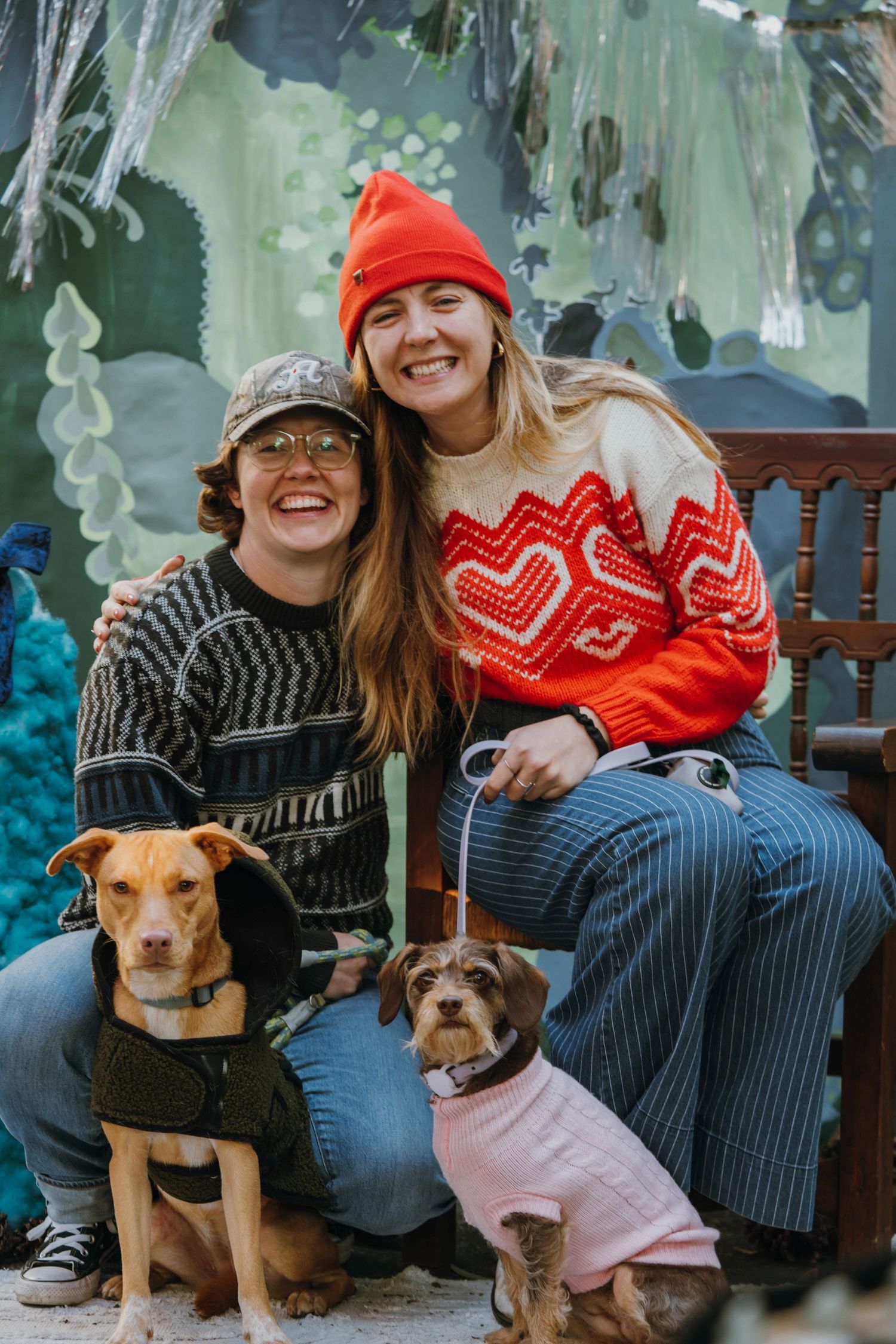 Two smiling women pose with two dogs in sweaters outdoors, one wearing a red beanie and heart sweater, the other in a patterned pullover.