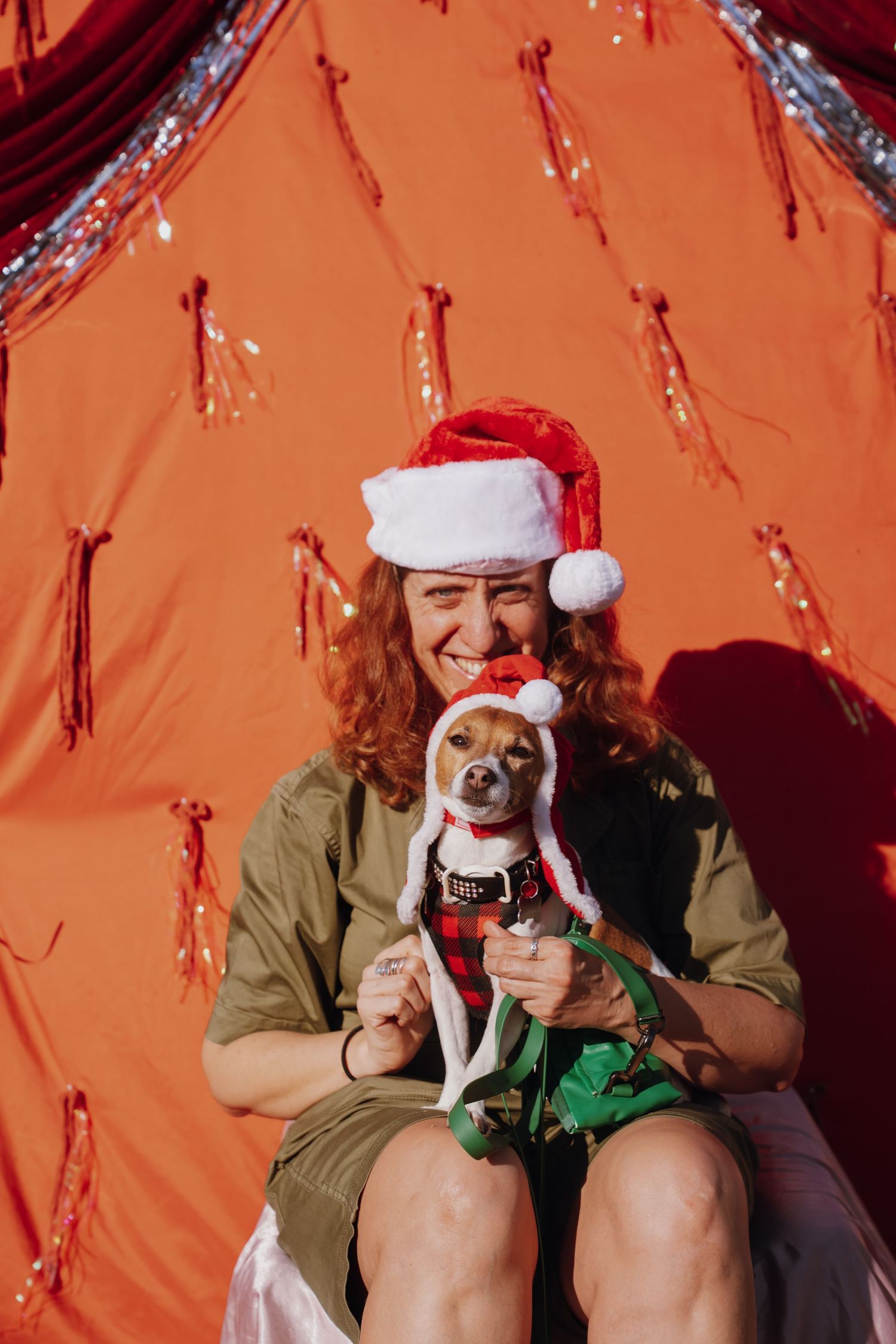 Woman in Santa hat laughing while holding a small dog dressed in an elf costume against a festive red background.