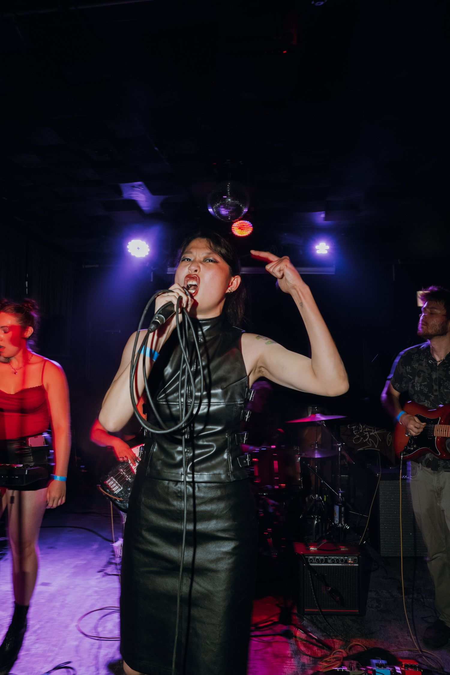 Female vocalist in black leather dress performing energetically on stage with band under purple lights.