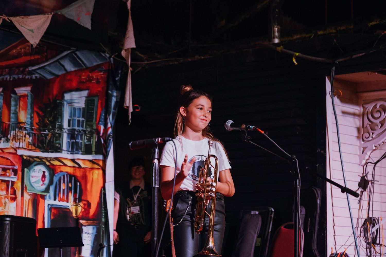 Smiling young girl holding a saxophone on a colorful painted stage at a music event.