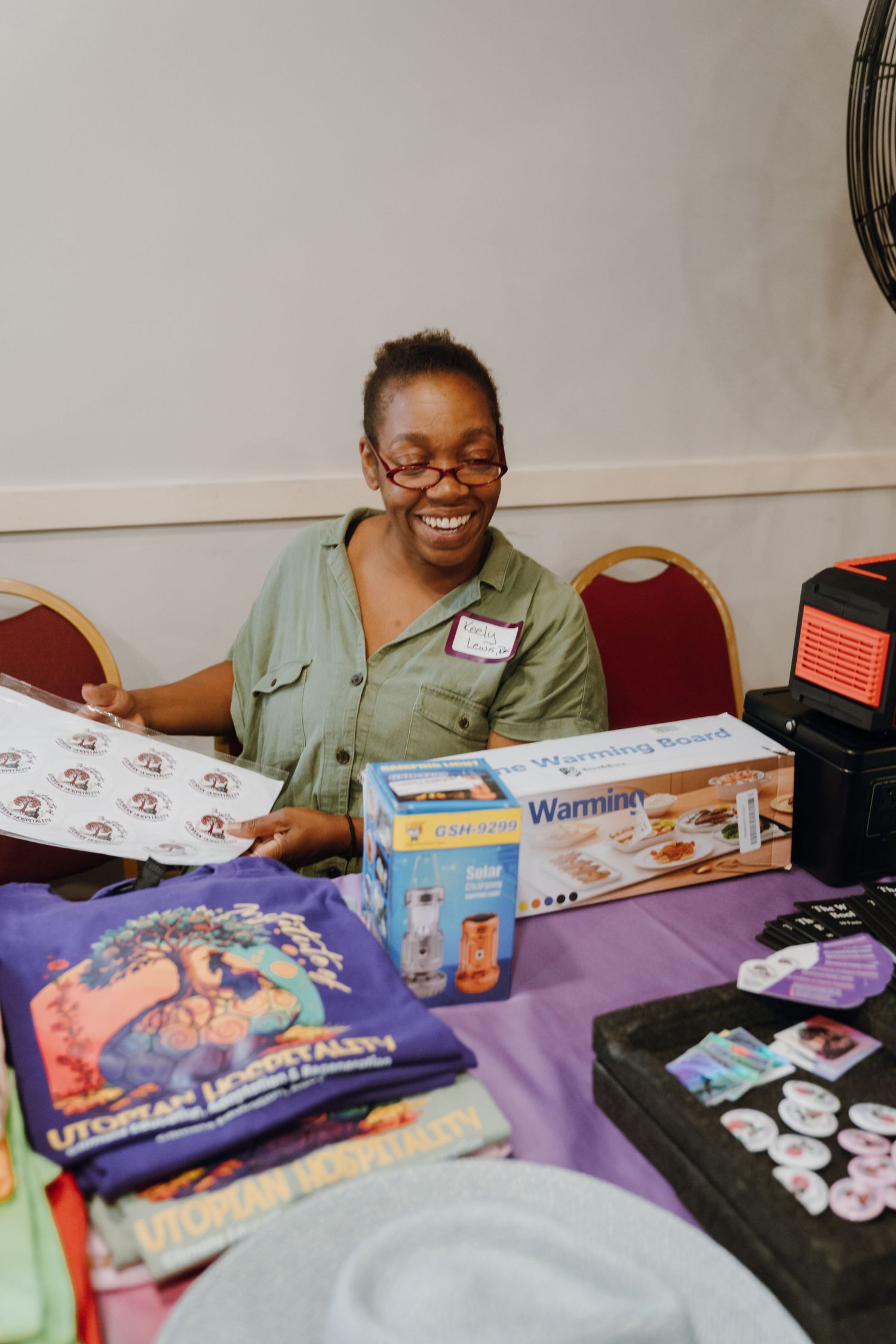 Smiling woman at a vendor table displaying colorful merchandise and products indoors.