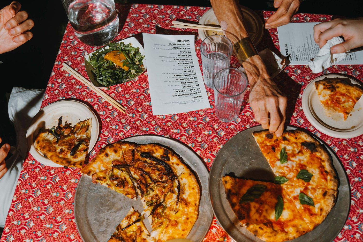Group of people sharing multiple pizzas on a red patterned tablecloth at a restaurant dinner.