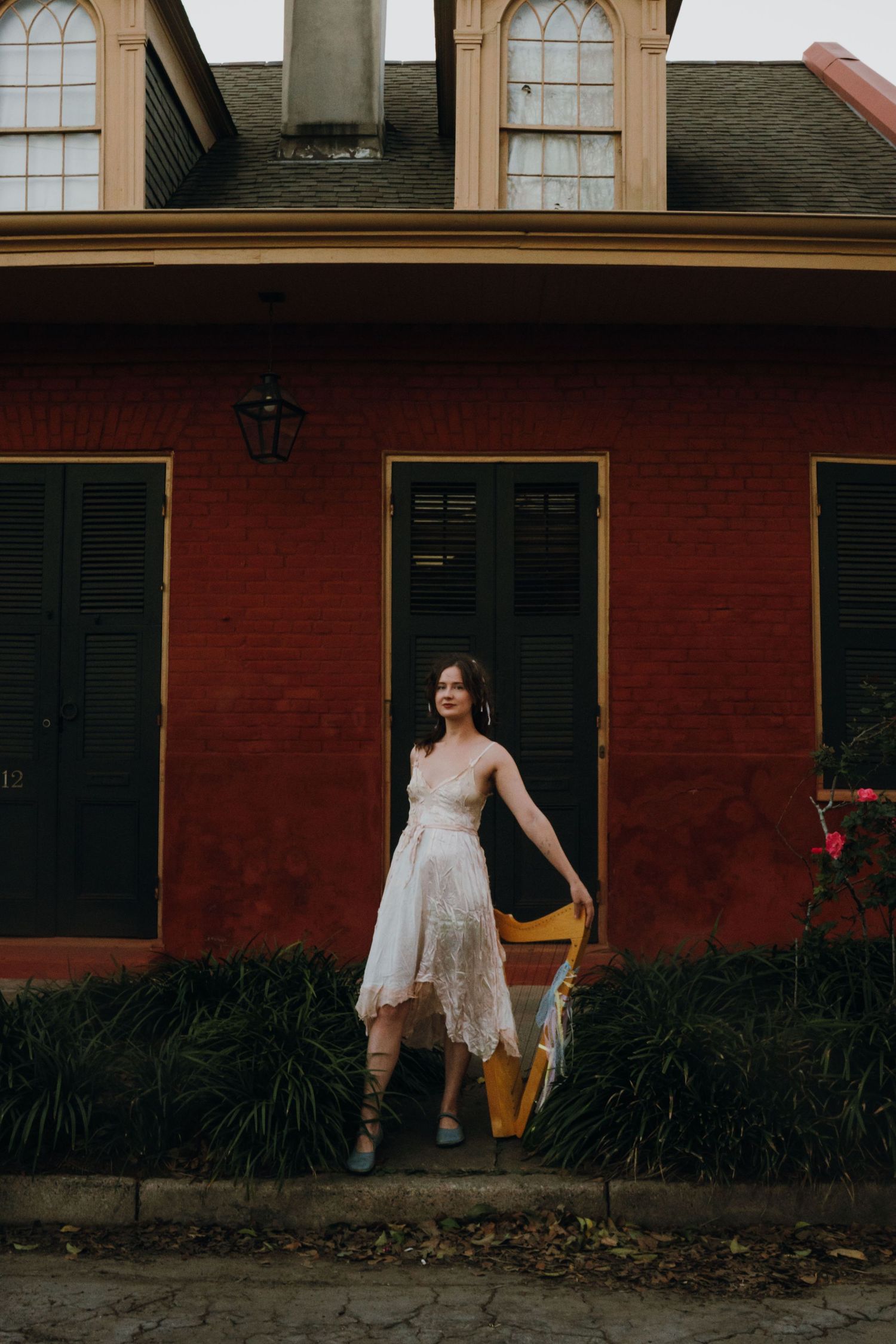 Woman in white lace dress holding a yellow harp stands before a red building with black shutters.