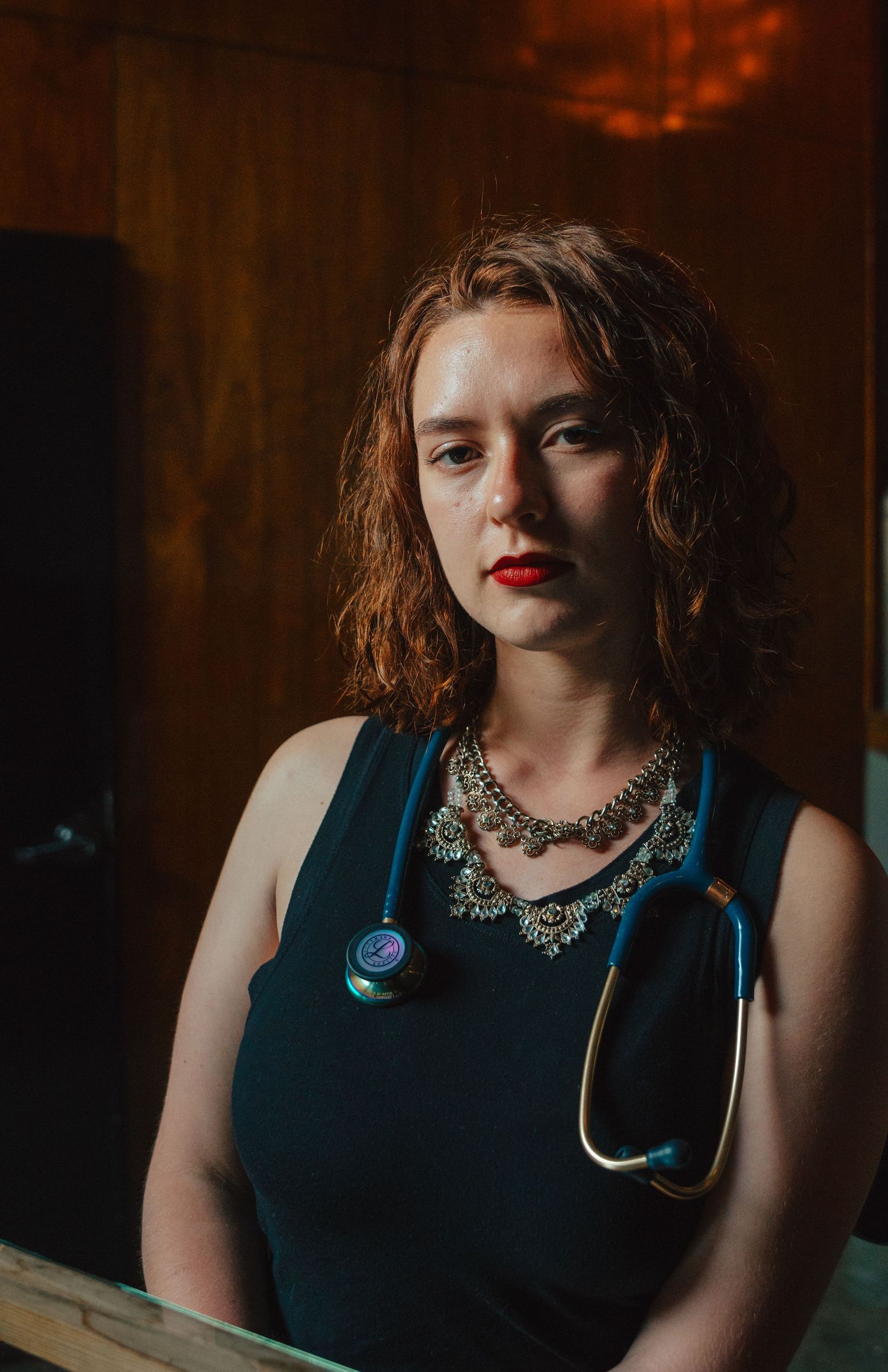 Young woman with red hair and red lipstick wearing teal dress with statement necklace and stethoscope.