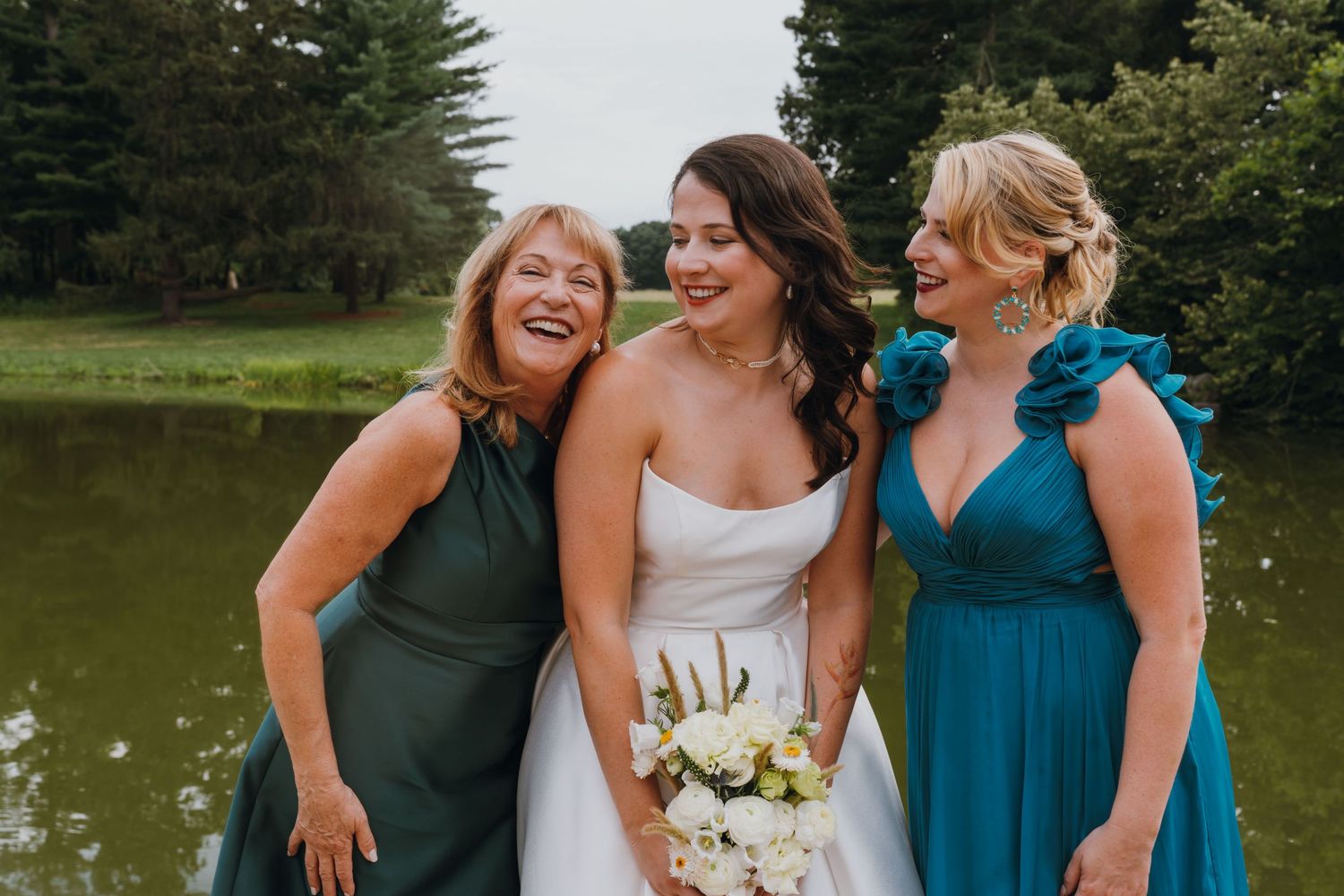 Bride in white strapless gown laughs joyfully with two bridesmaids in teal and green dresses by a pond. New Orleans, Louisiana, Mississippi