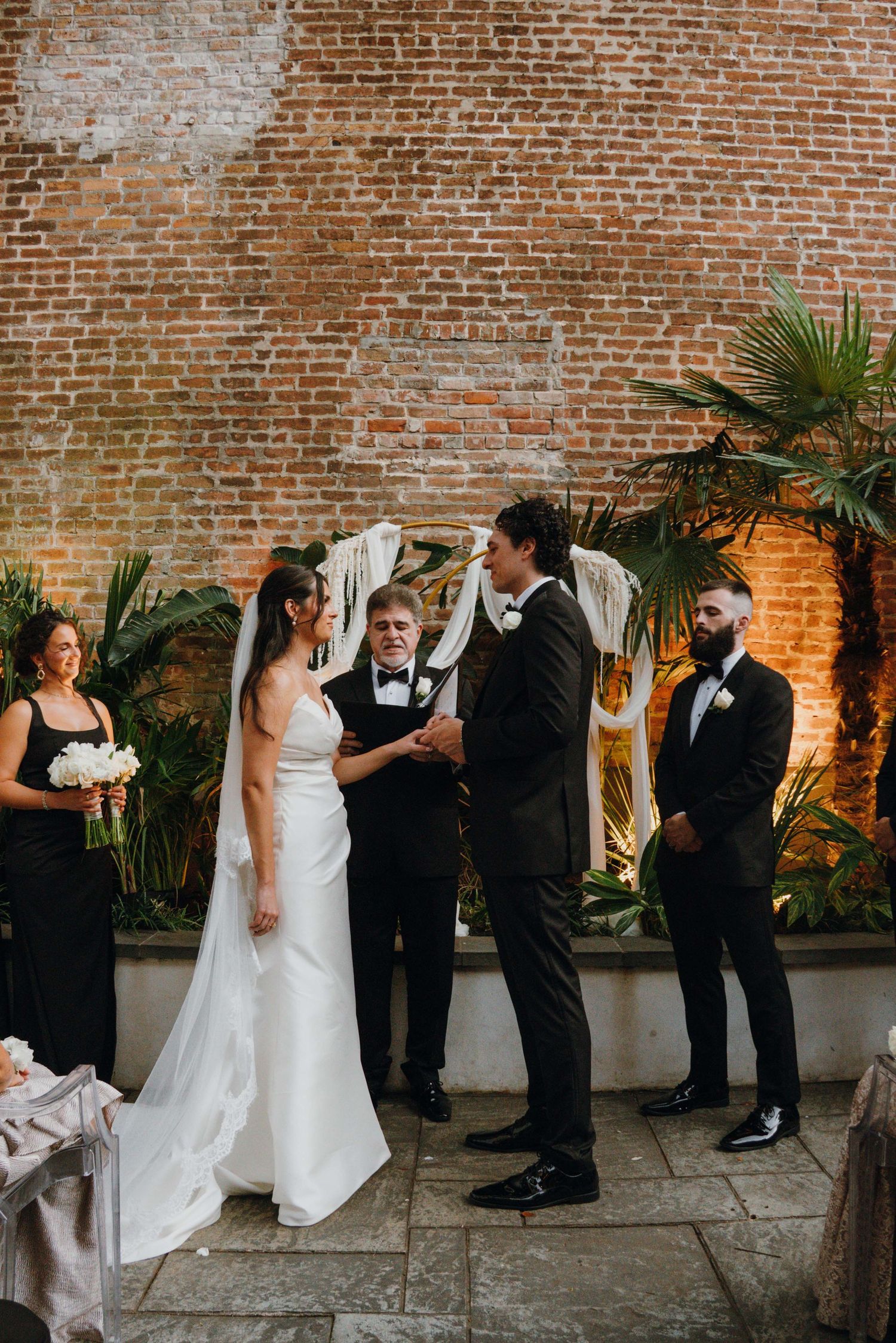 Bride and groom holding hands at outdoor wedding ceremony with brick wall and tropical greenery backdrop.