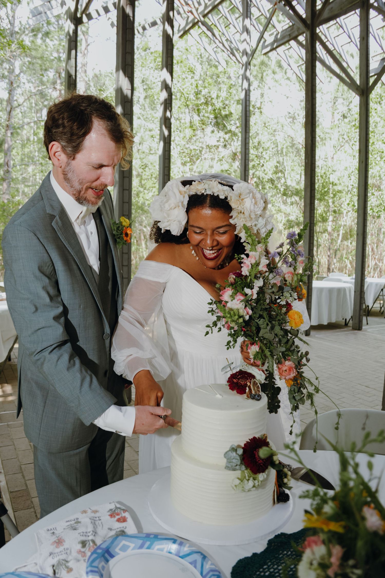 Interracial couple joyfully cuts white tiered wedding cake adorned with fresh flowers.