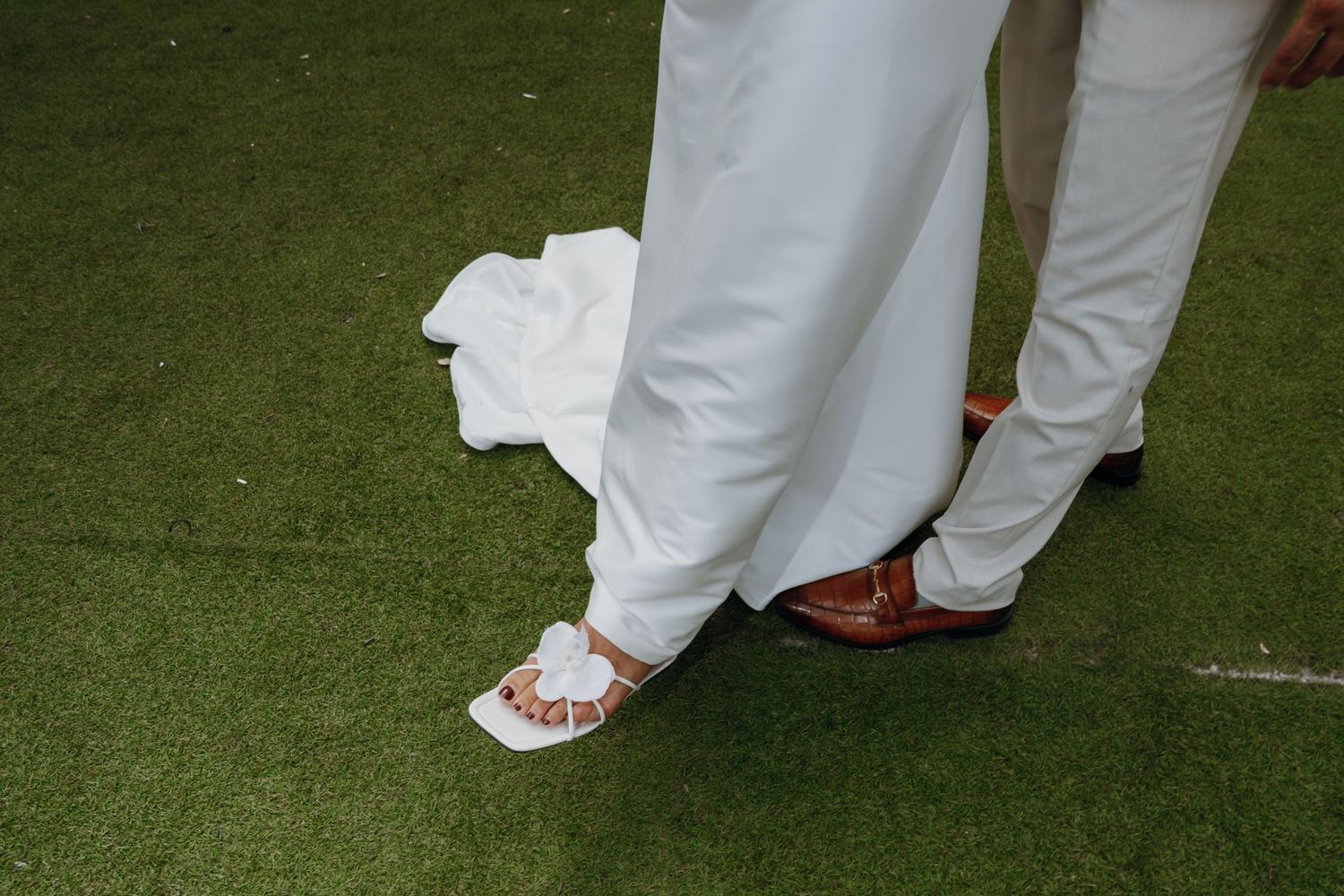 Close-up of bride in white sandals and groom in brown leather shoes standing on green turf.