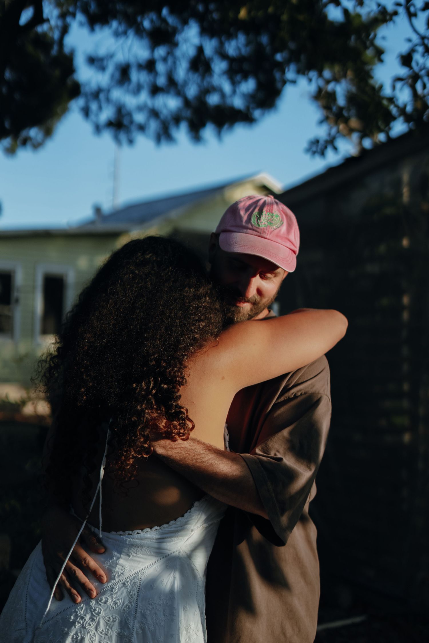 A couple shares a warm embrace outdoors in golden sunlight beneath a large tree.