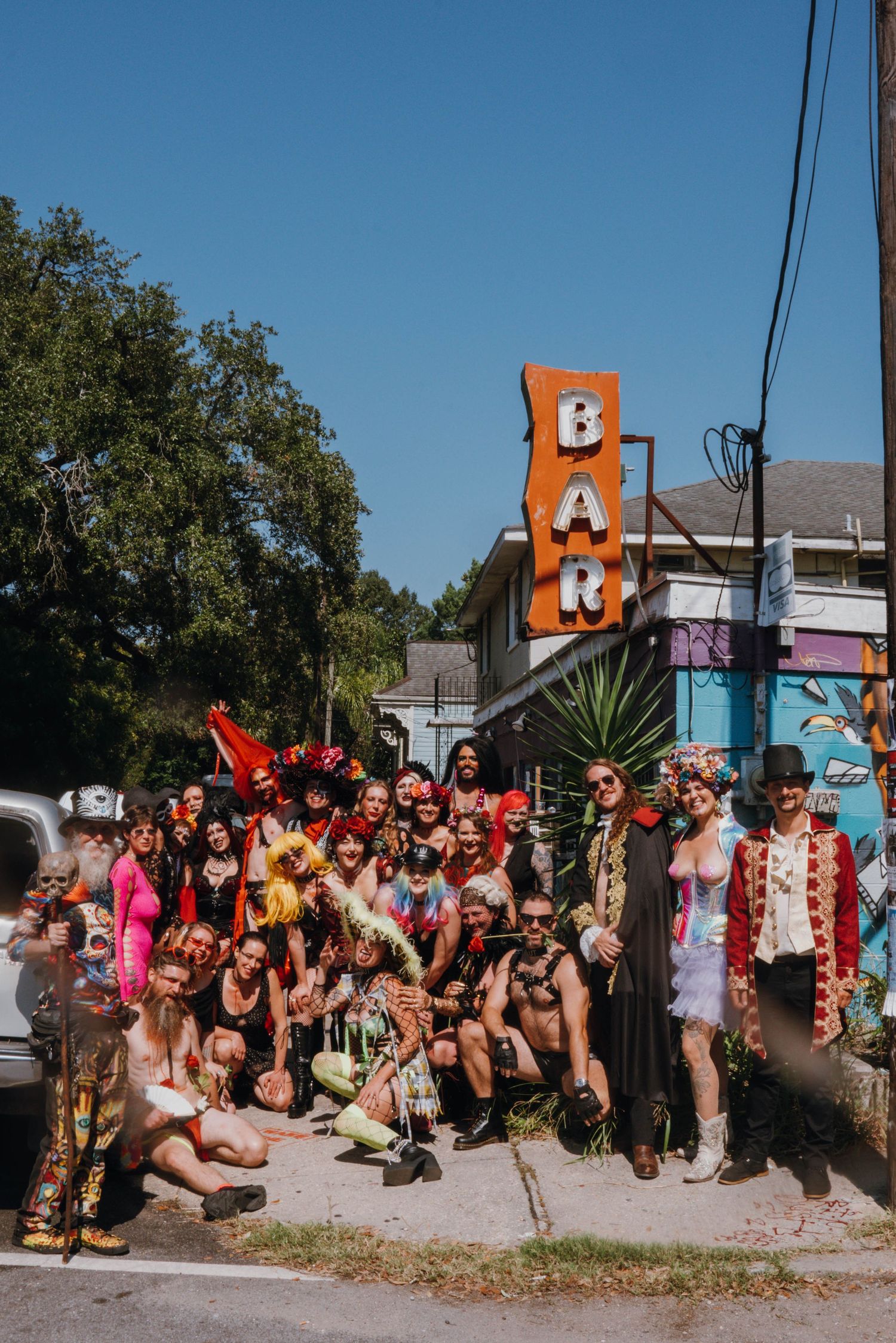 Large group of elaborately costumed wedding guests posing outside Bar sign on a sunny day.