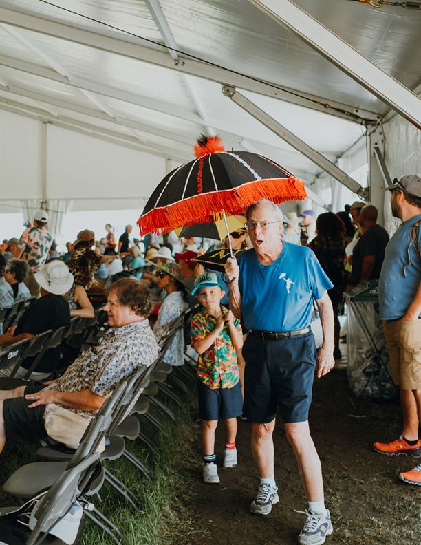 Elderly man holding decorative umbrella at outdoor festival event under white tent with crowd seated.