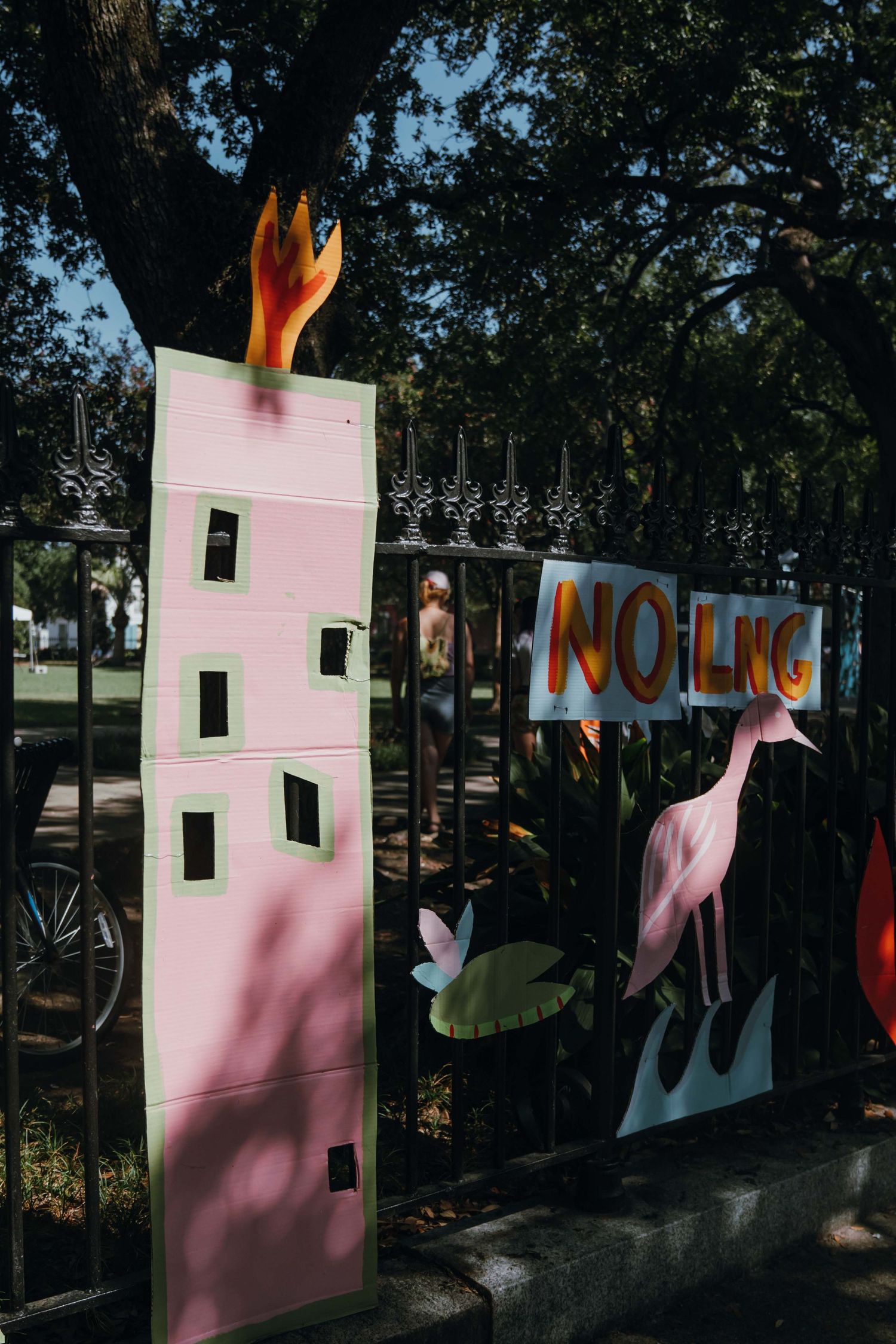 Colorful outdoor protest art display with NO LNG sign and pink building cutout on iron fence under trees.
