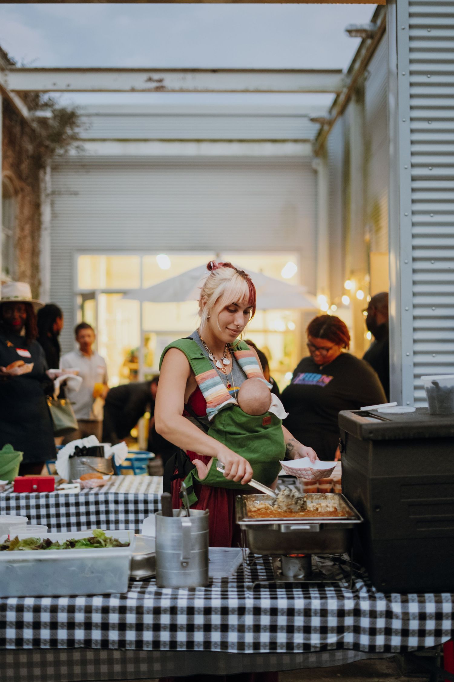 Woman carrying baby while serving food at an outdoor community gathering buffet table.