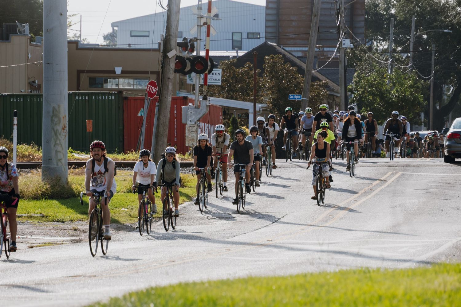 Large group of cyclists riding together down a road near a railway crossing on a sunny day.