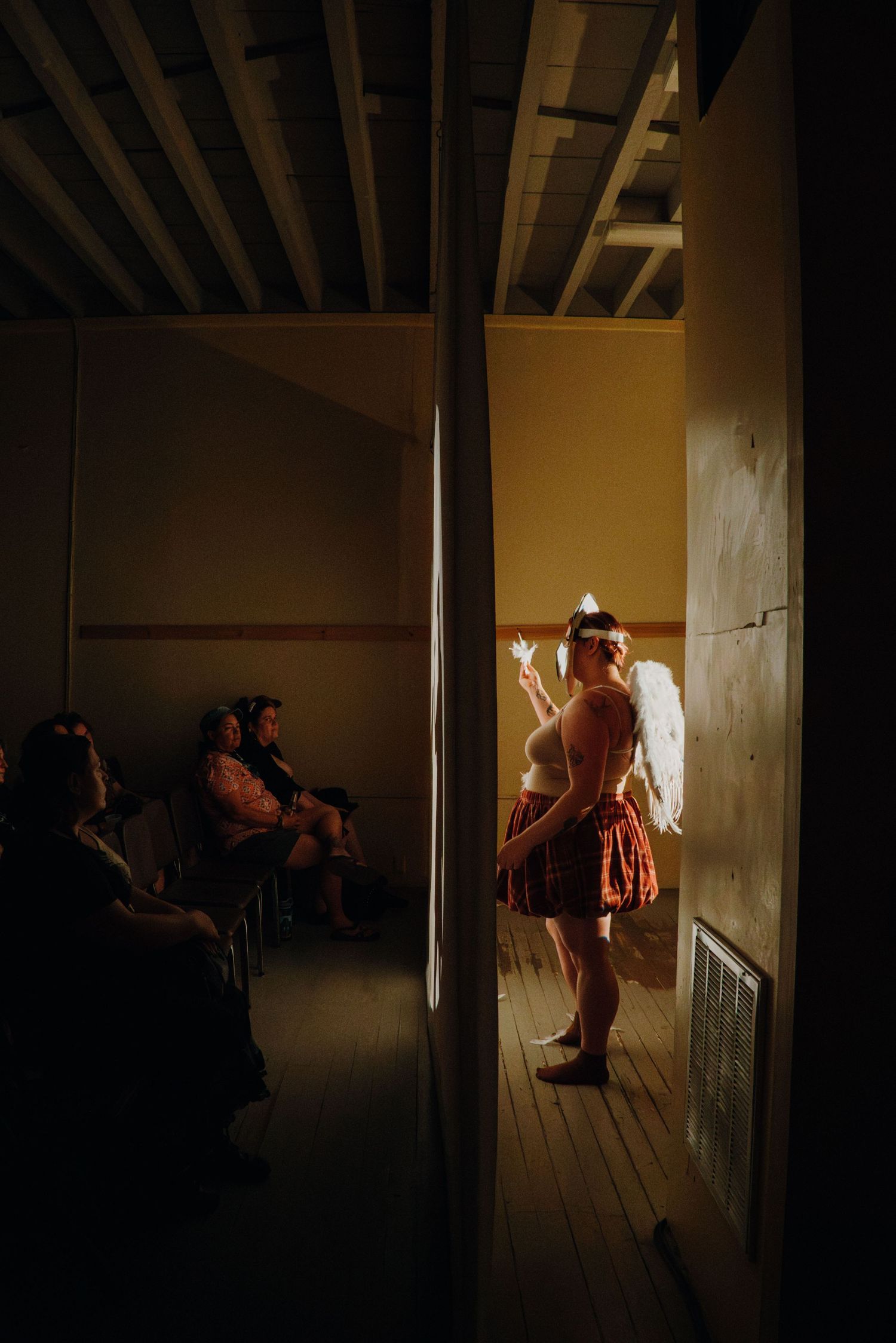 Angel-costumed performer stands in doorway as seated audience watches in dim room.