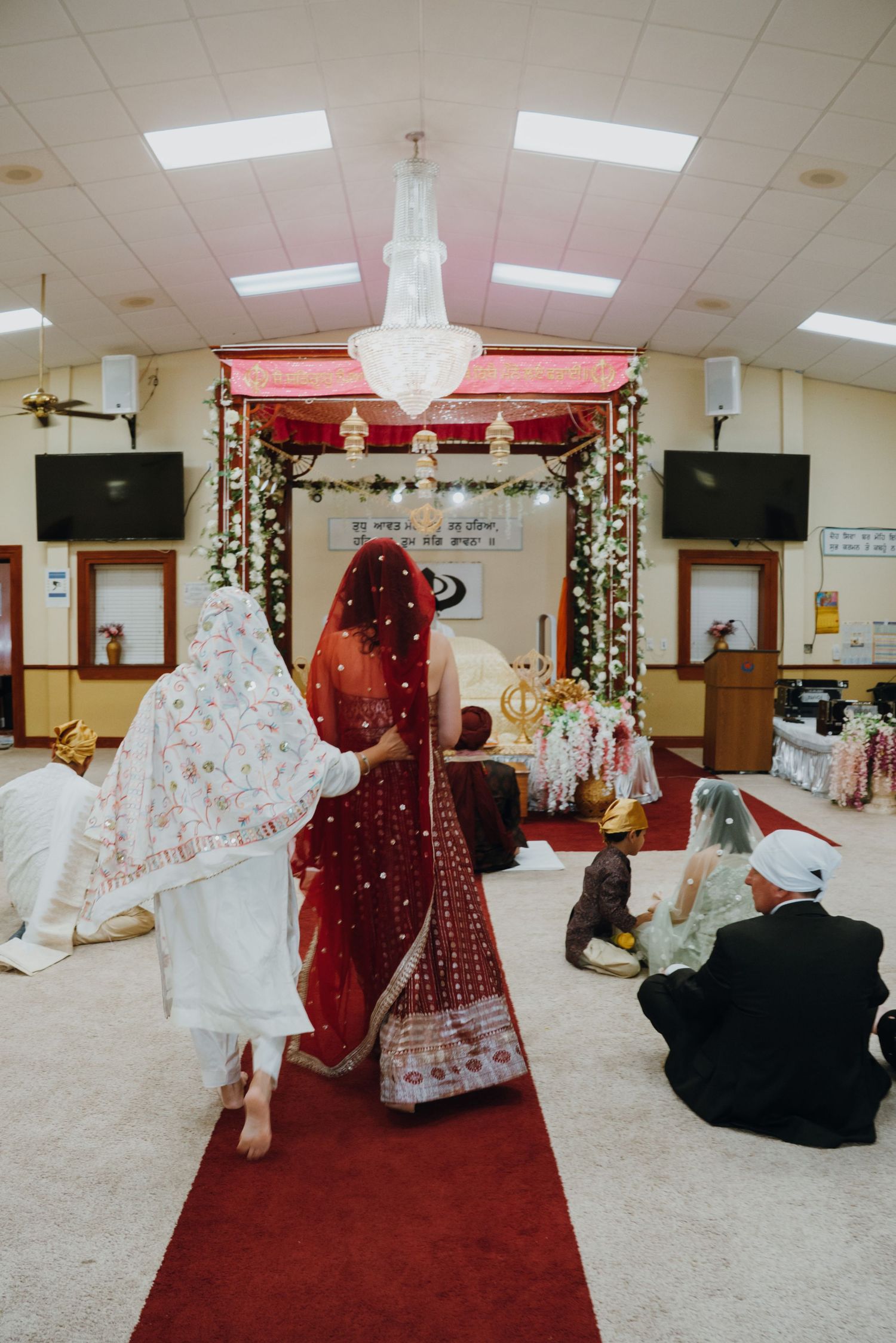 Sikh bride in red lehenga walks down the aisle toward the decorated altar during Anand Karaj ceremony.