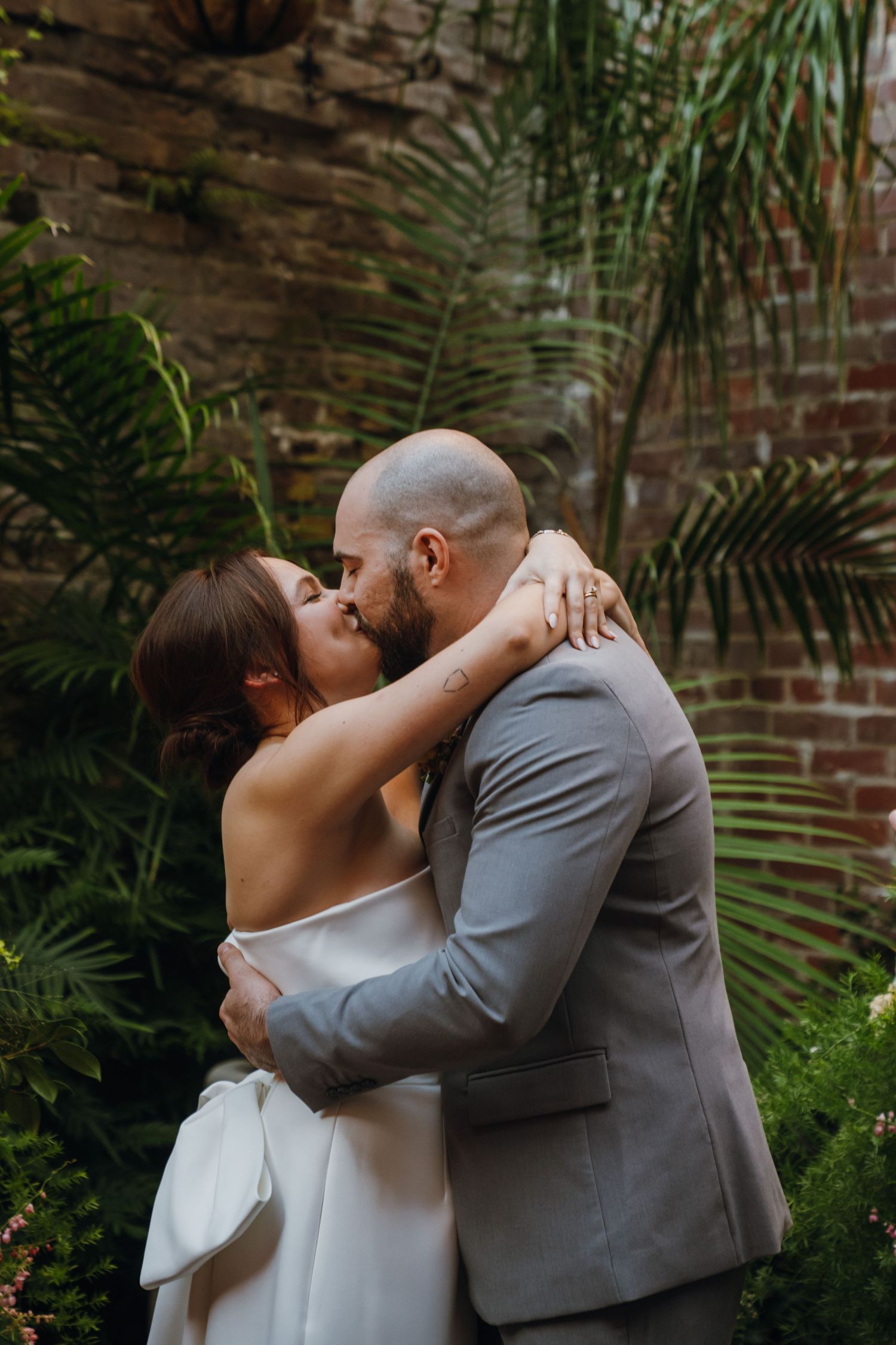 Bride and groom share a romantic first kiss surrounded by lush tropical greenery and rustic brick wall. New Orleans, Louisiana, Mississippi