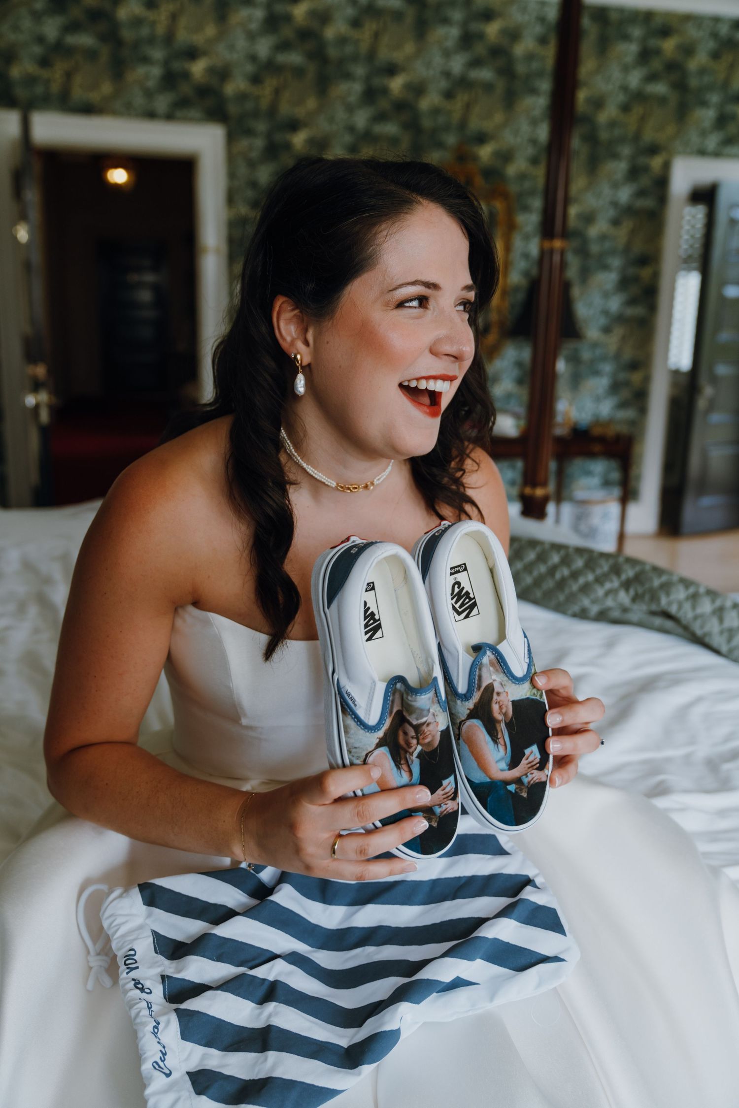 Excited bride in white strapless gown holding custom photo-printed Vans sneakers while sitting on a bed. New Orleans, Louisiana, Mississippi