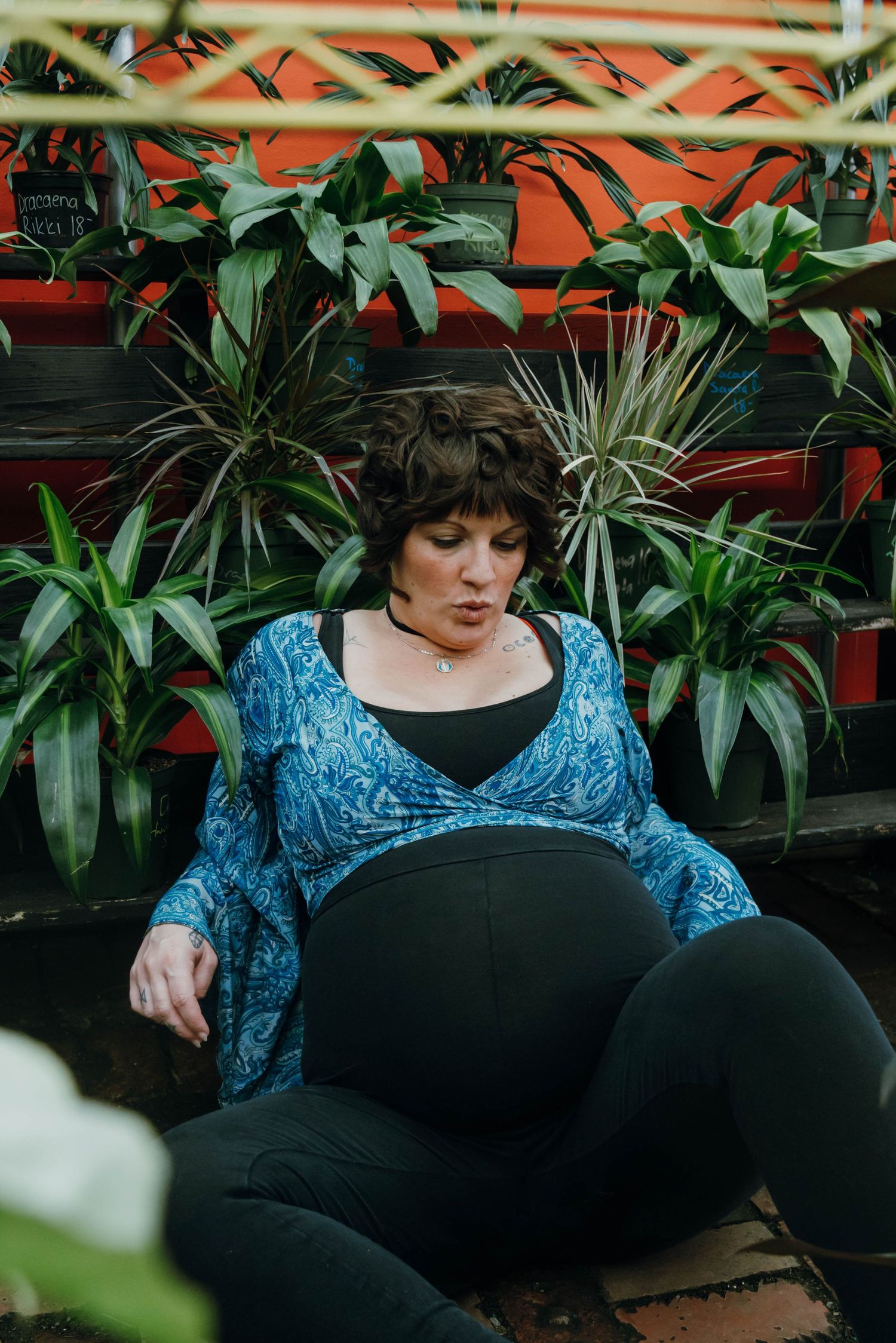 Pregnant woman in blue paisley cardigan sitting among tropical plants during outdoor maternity shoot.