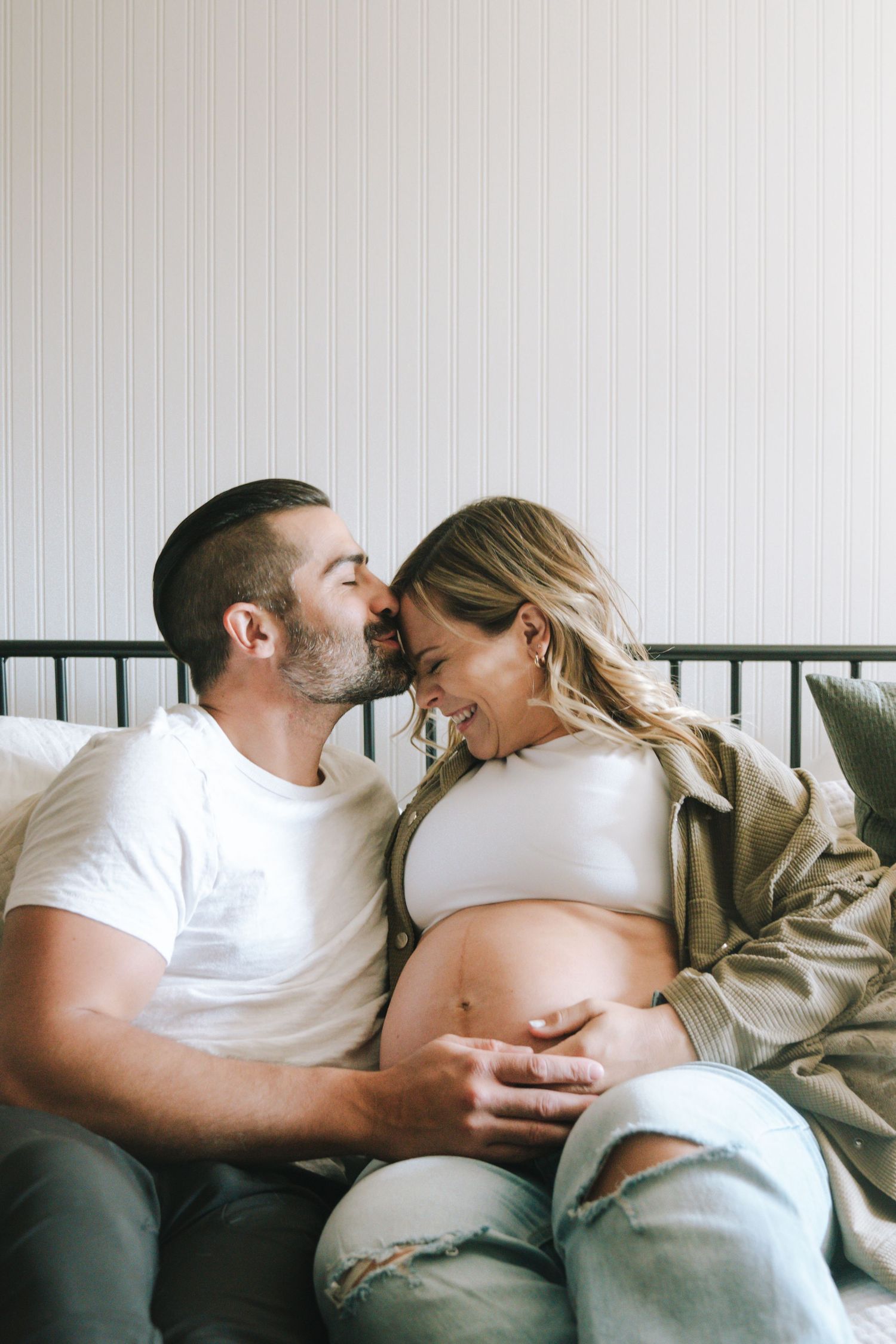 Happy couple on bed, man kissing pregnant woman's head while holding her bump.