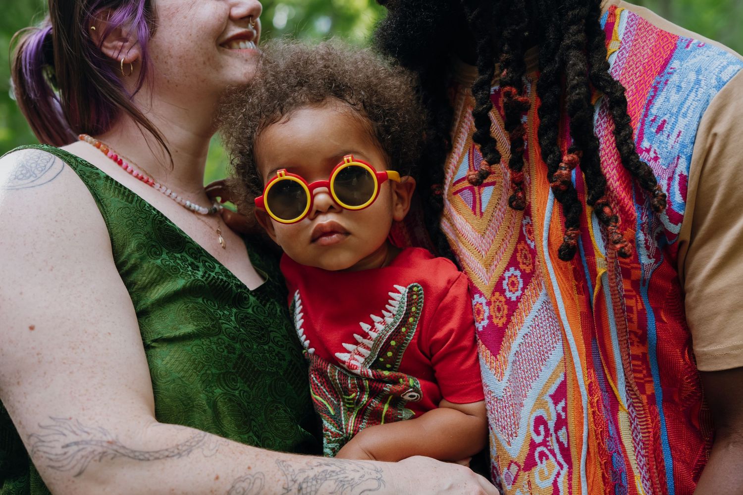 Adorable toddler in red outfit wearing yellow round sunglasses held by two parents outdoors.