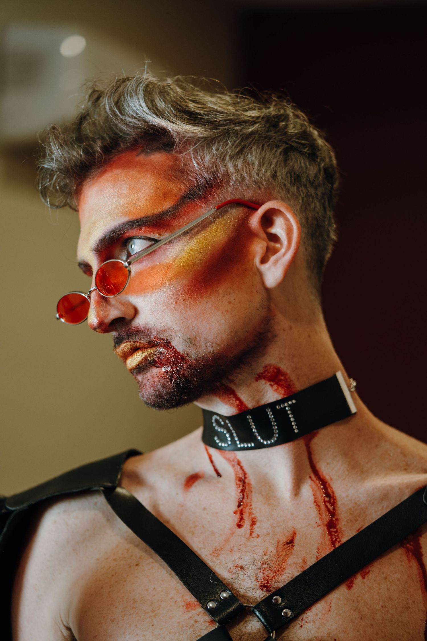 Man with dramatic red and orange face paint wearing round red sunglasses and a leather choker collar.