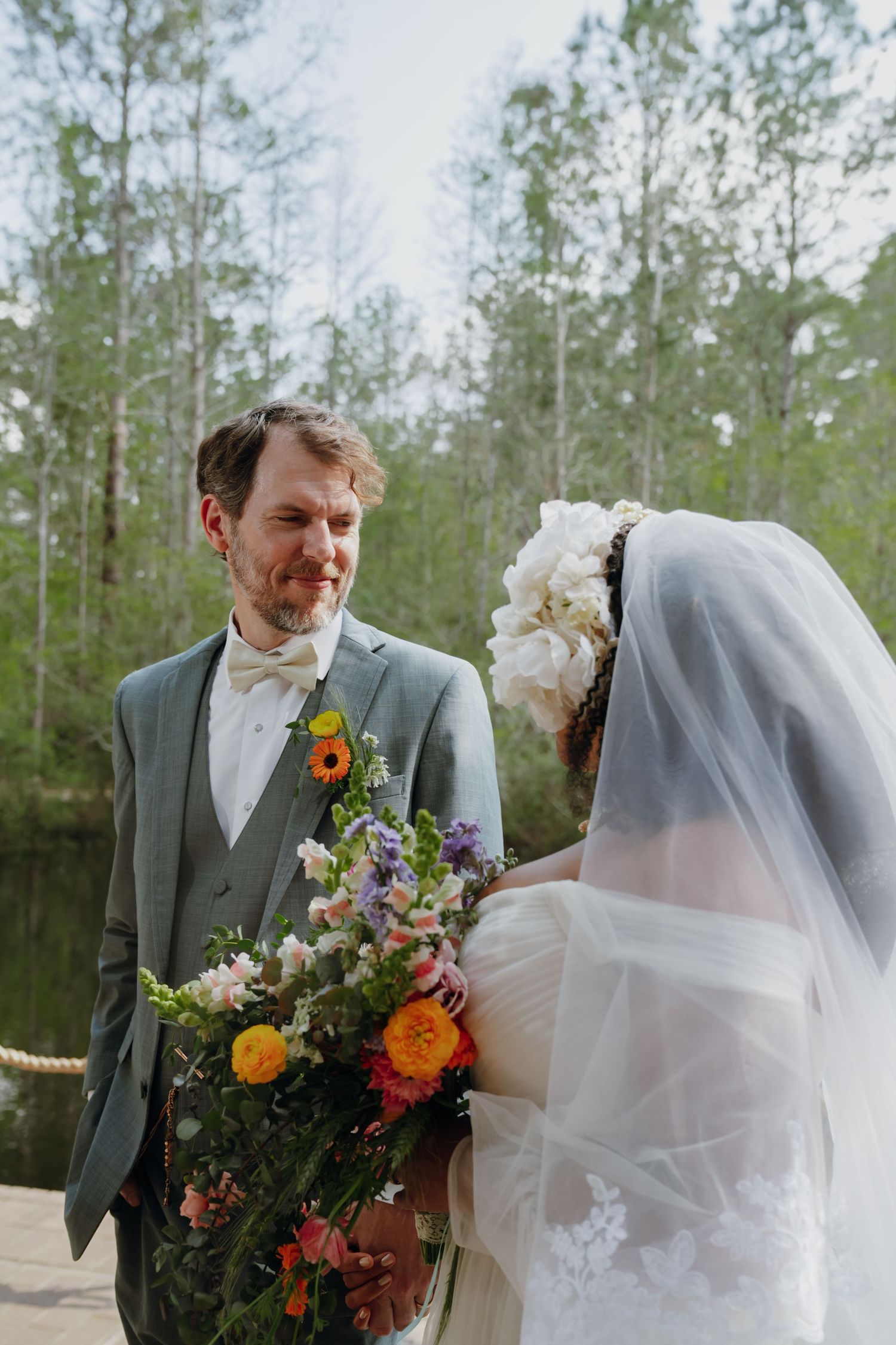 Groom smiles lovingly at bride holding vibrant wildflower bouquet by lakeside.