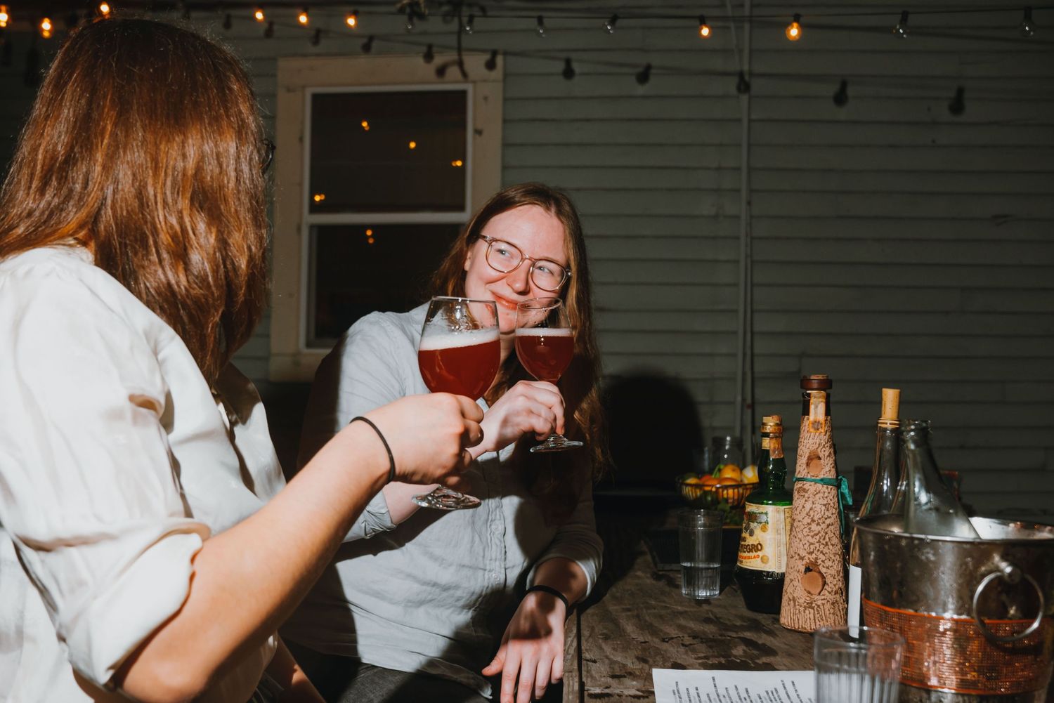 Two women toasting with craft beer glasses and smiling at an outdoor evening gathering.
