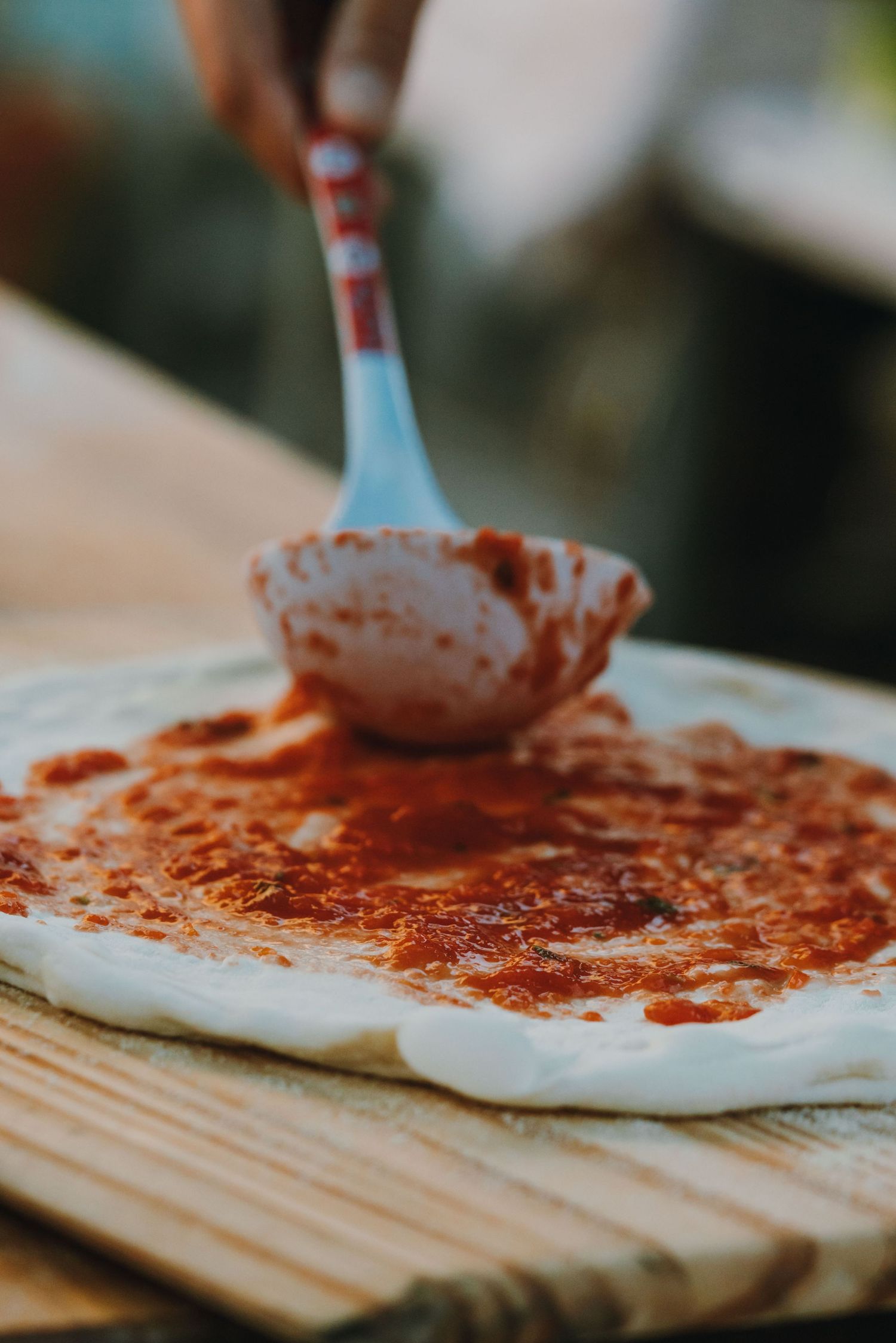 Hand spreading tomato sauce on fresh pizza dough with a ladle on a wooden board.