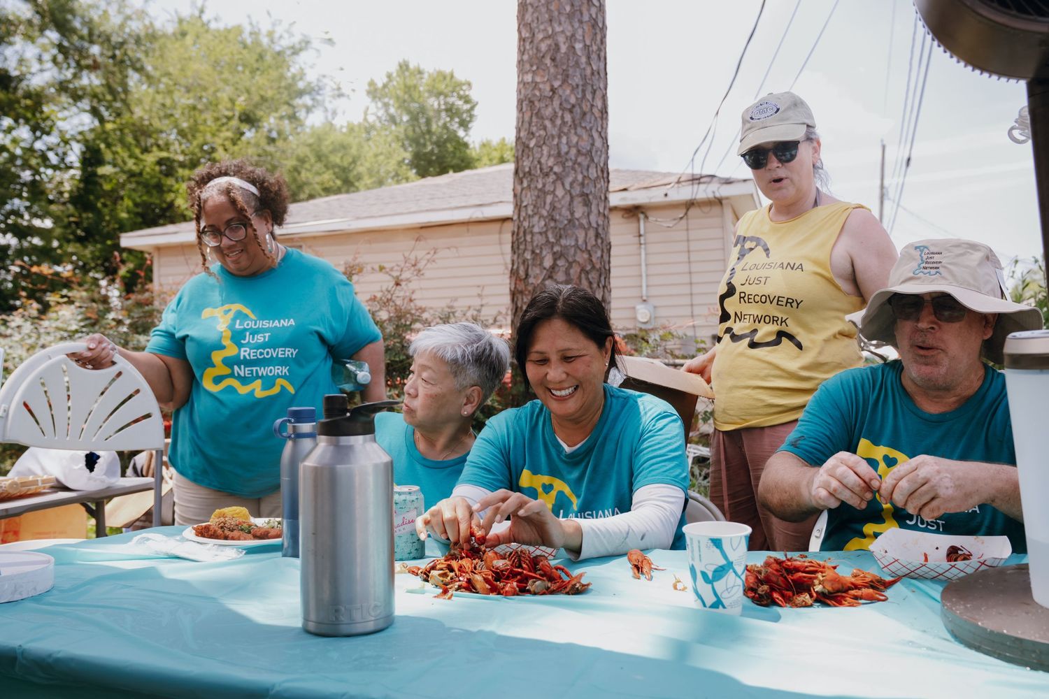 Group of volunteers in teal shirts enjoying a meal of crawfish together at an outdoor picnic table.