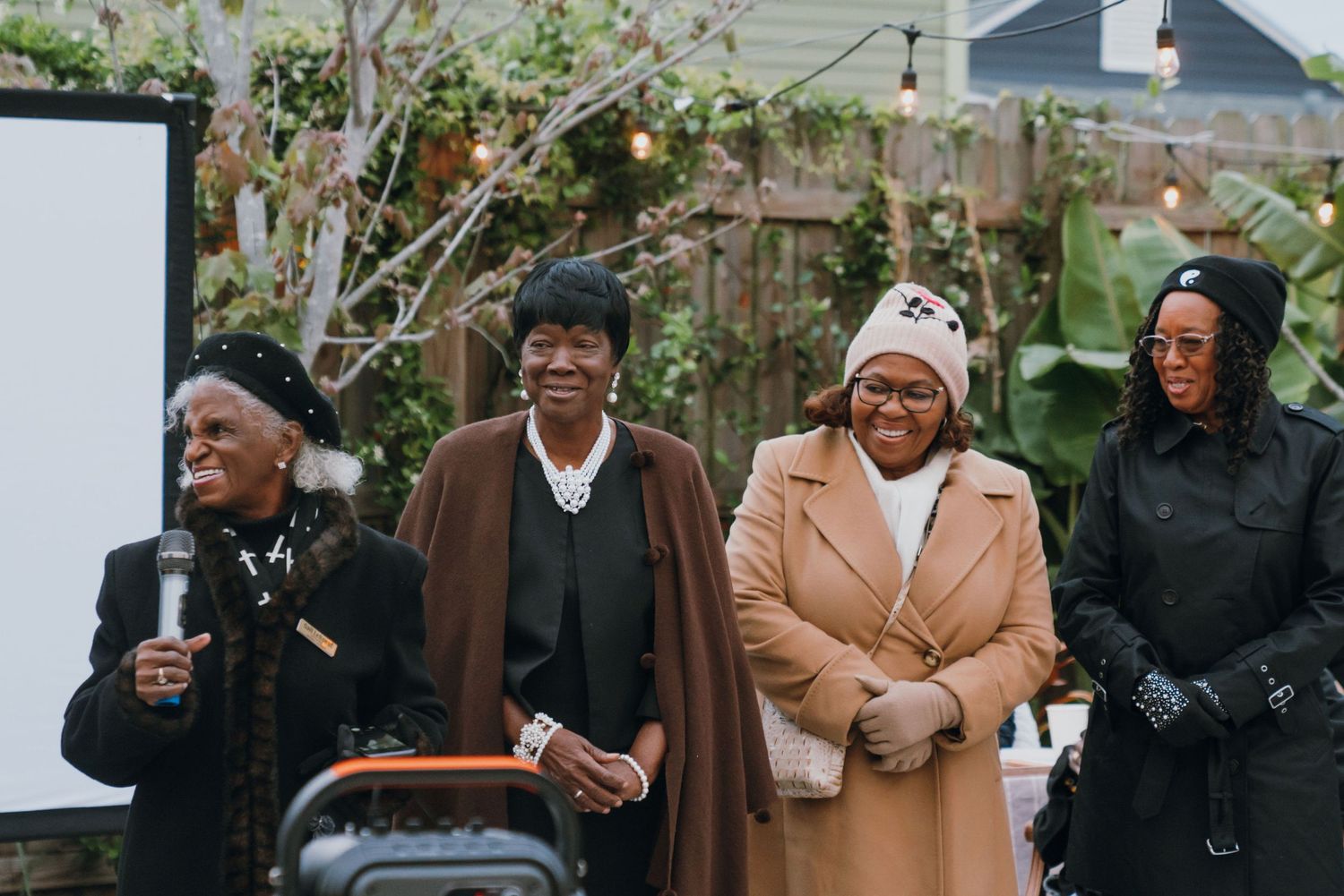 Four women smiling outdoors at a community event with string lights and a projector screen.